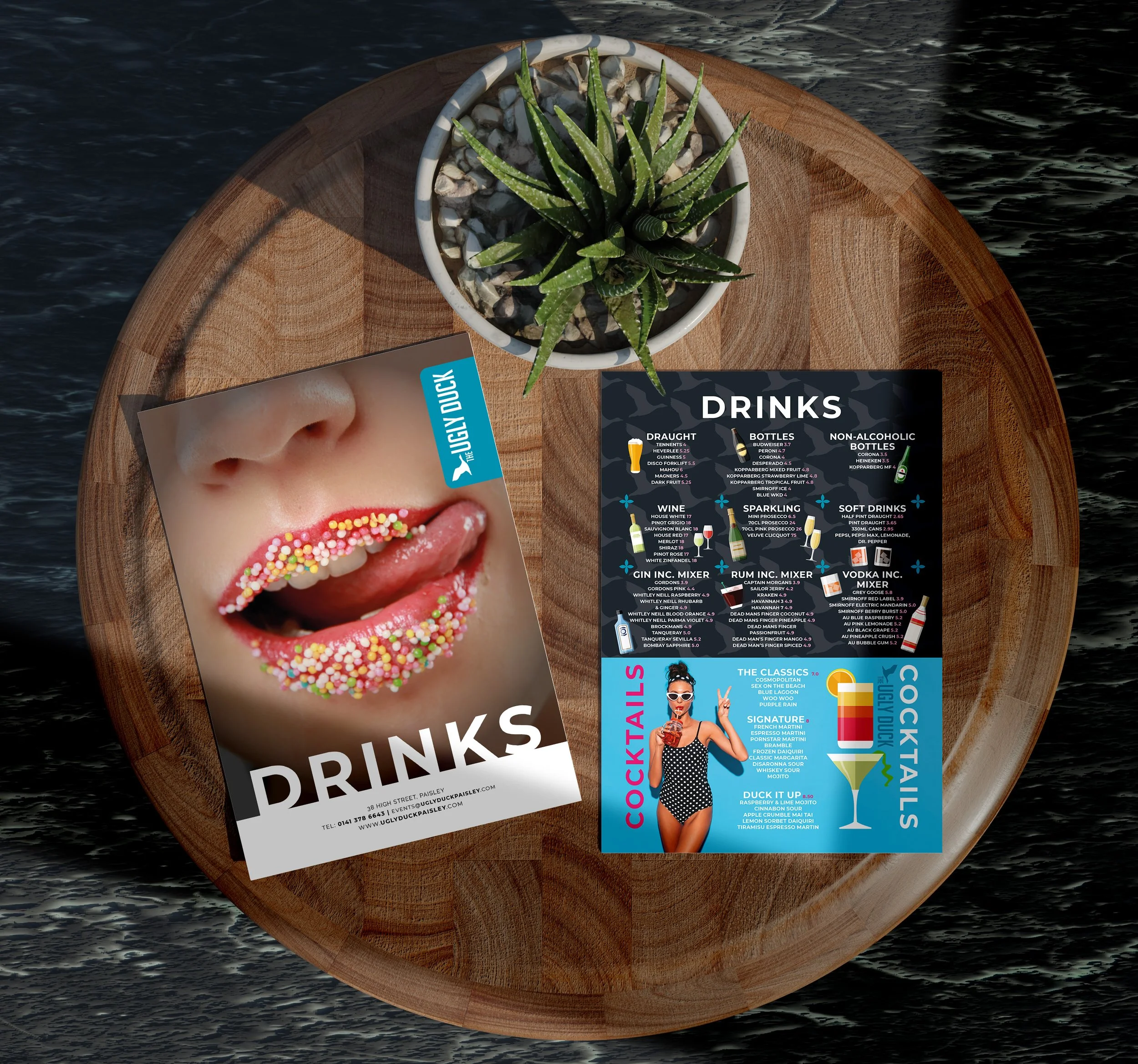 A round wooden table with a small potted succulent plant and two cocktail menus, one featuring a woman with sprinkles on her lips and the other listing various drink options.