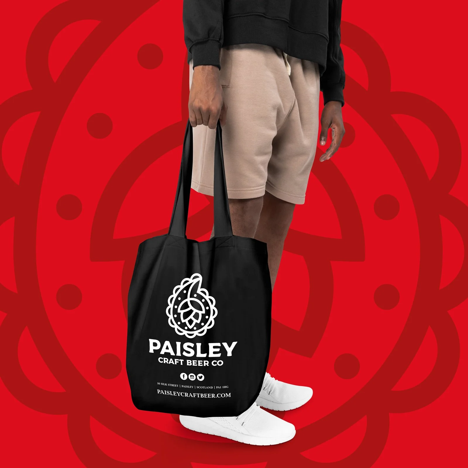 A person wearing beige shorts and white sneakers holding a black tote bag with a Paisley craft beer logo, standing against a red background with a Paisley pattern.