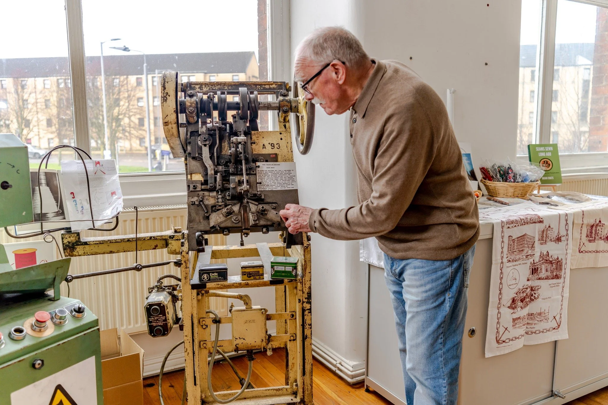 An elderly man with glasses working on an old industrial machine indoors near a window with a city view.