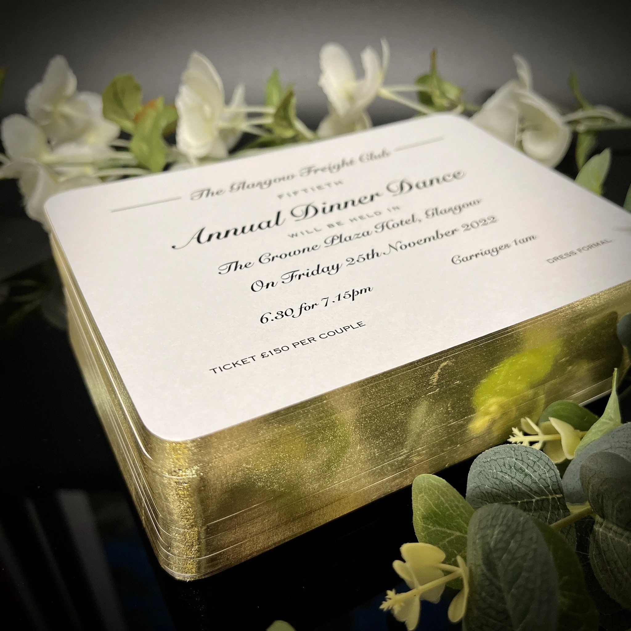 Invitation card for the annual dinner dance at The Groune Plaza Hotel, Glasgow, on November 25, 2022, from 6:30 to 7:15 pm, ticket costing £150 per couple, surrounded by white flowers and green foliage.