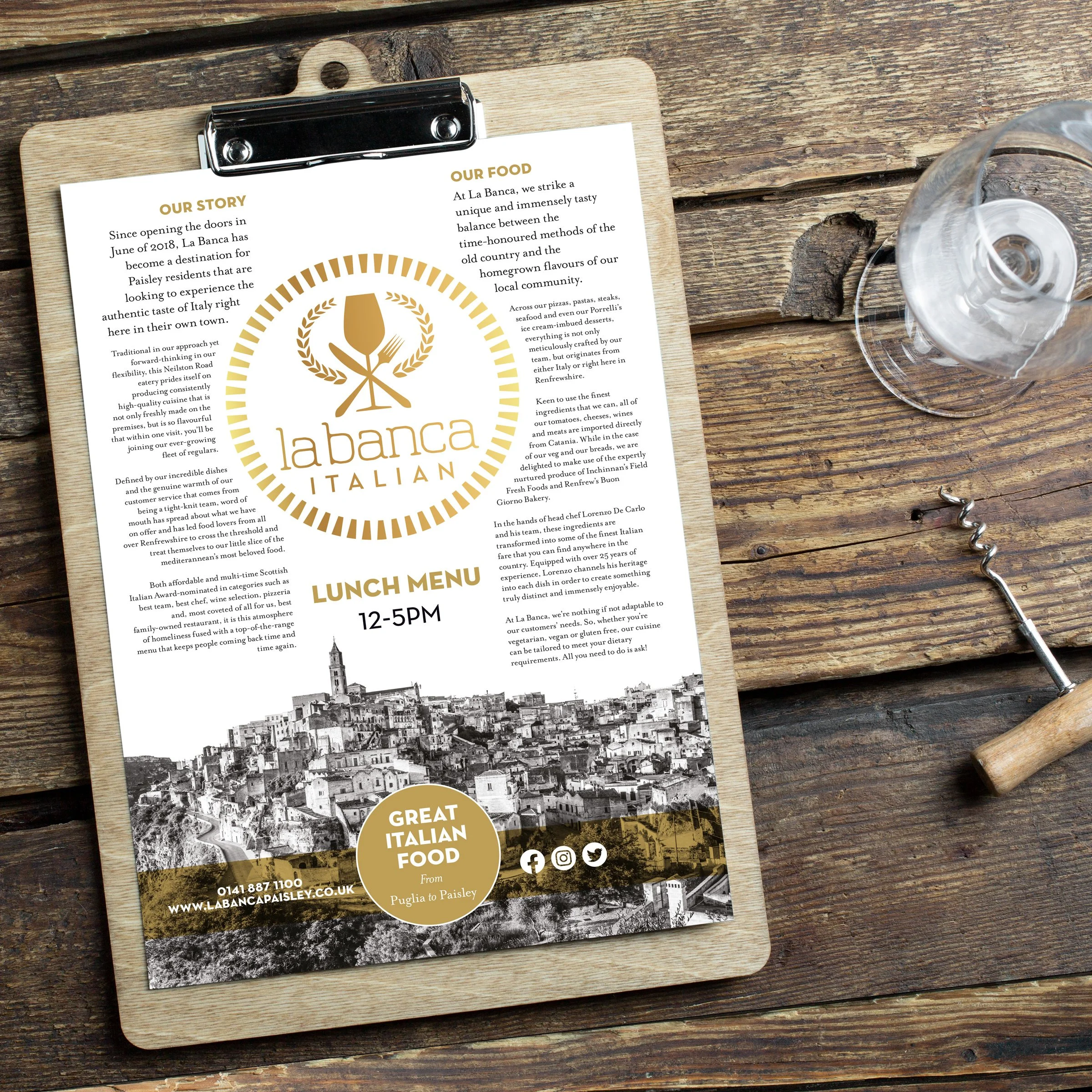 Menu on a clipboard for La Banca Italian restaurant, displaying lunch hours from 12 to 5 PM, with a black and white image of an Italian hillside town at the bottom and social media icons. The menu is on a wooden table with a glass of water and a cork