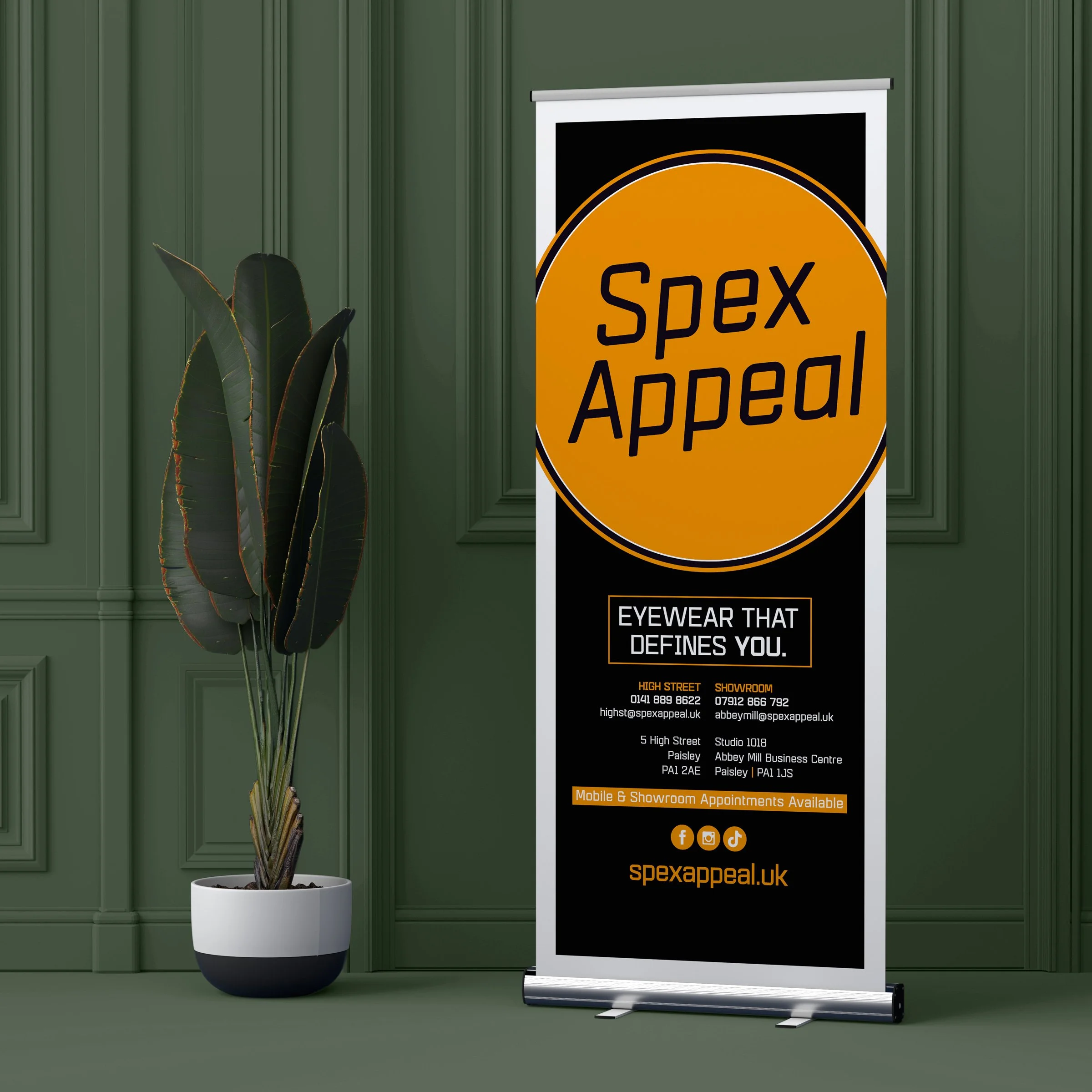 A vertical banner for Spex Appeal, an eyewear store, with a large orange circle and black background, displaying the store's contact information, address, and social media icons. The banner is placed next to a potted plant with dark green leaves in a