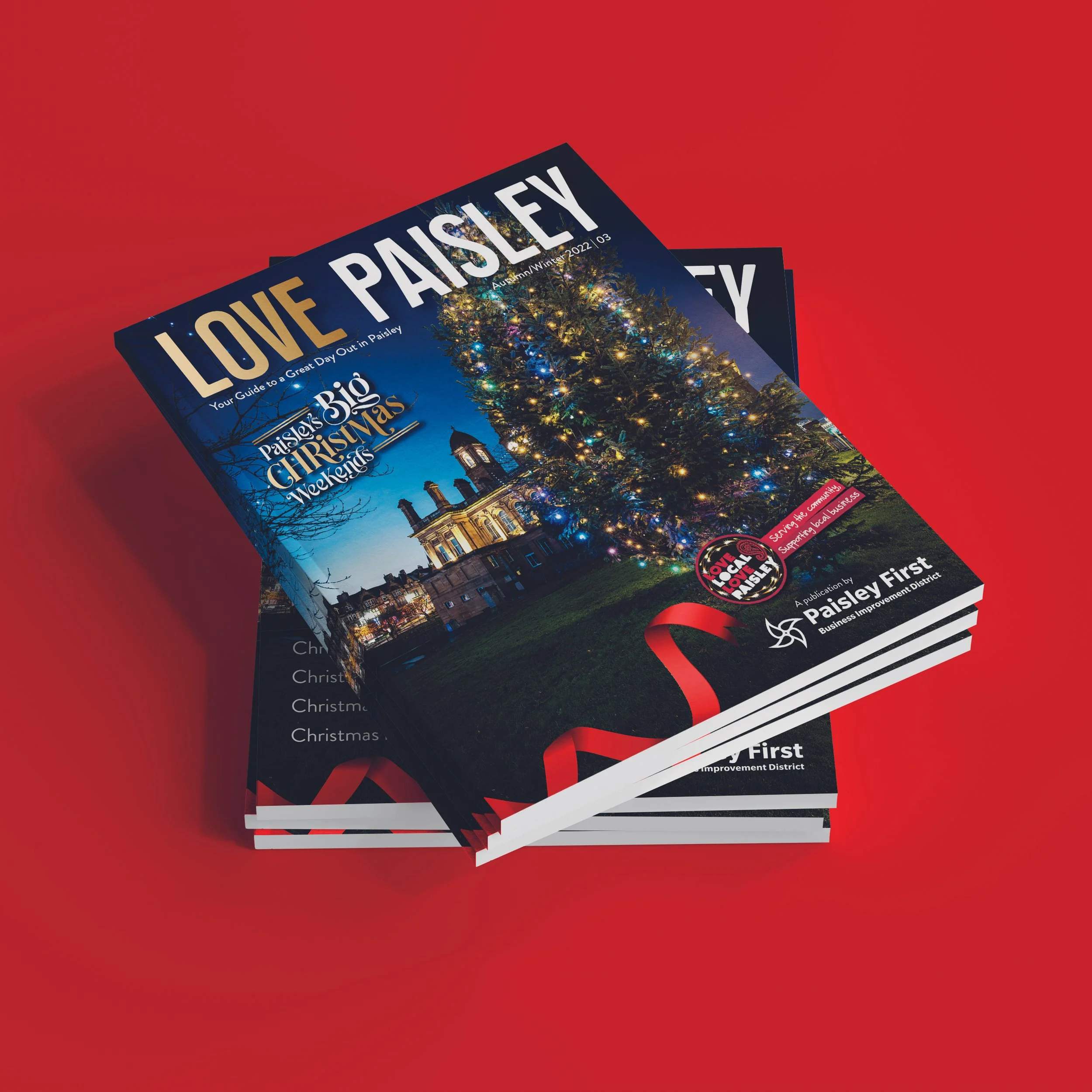 Stack of magazines titled 'Love Paisley,' featuring a Christmas tree decorated with lights and ornaments on the cover, with a historic building and a dark blue sky in the background.