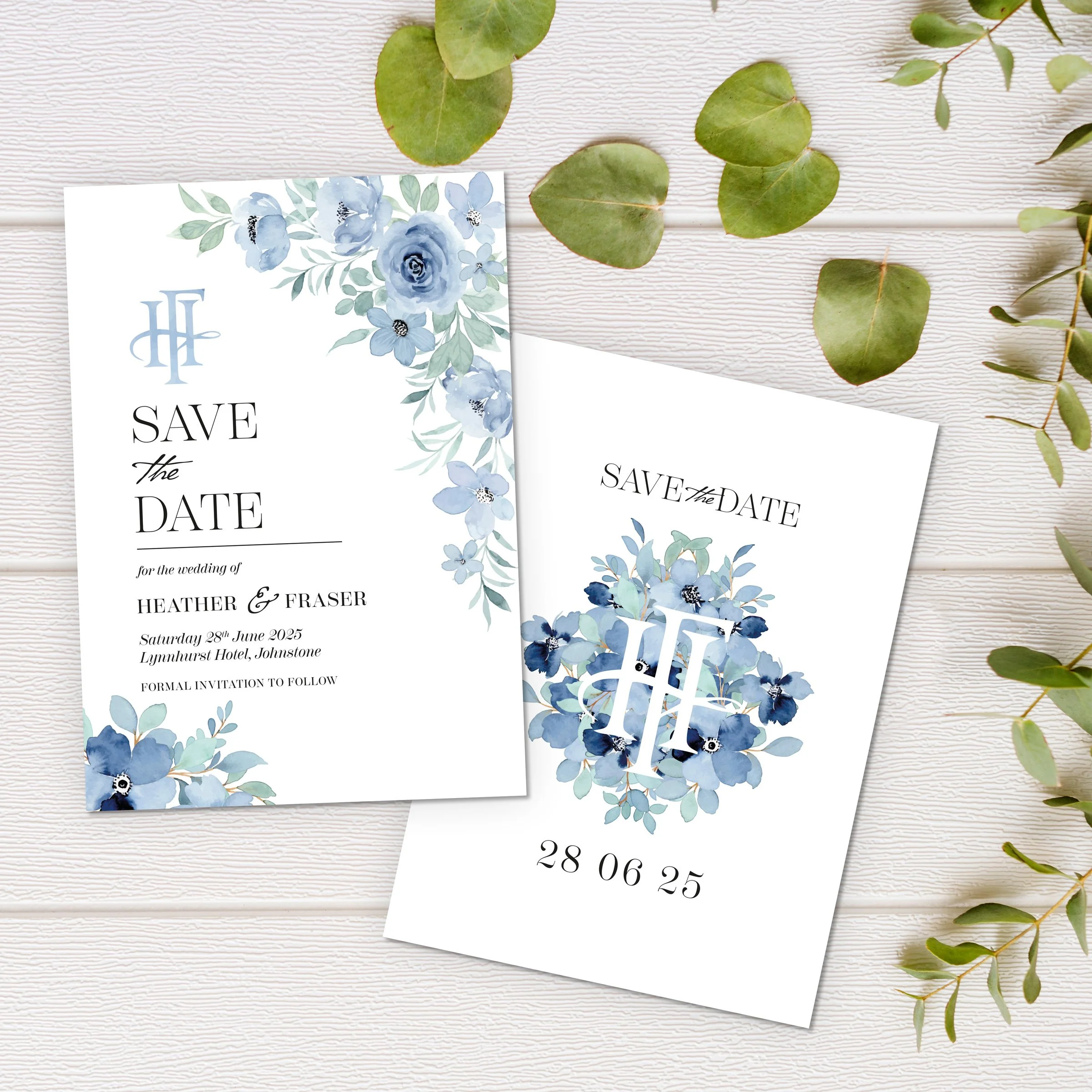 White wedding save the date card with blue floral accents and monogram, placed on white wooden surface with green leaves.