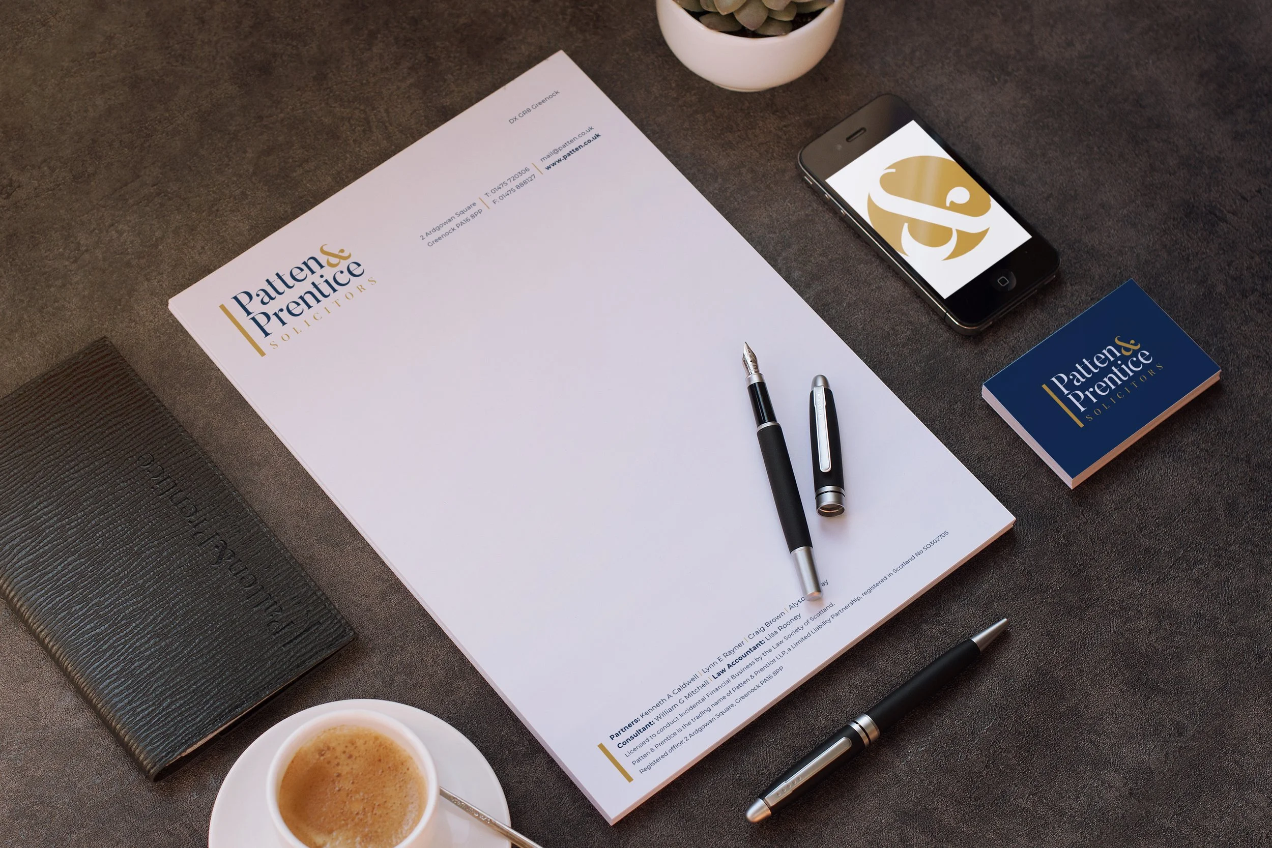 Business workspace with document labeled 'Patten & Prentice Solicitors', black notebook, cup of coffee, pens, a deck of cards with the same logo, and a smartphone on a dark desk.