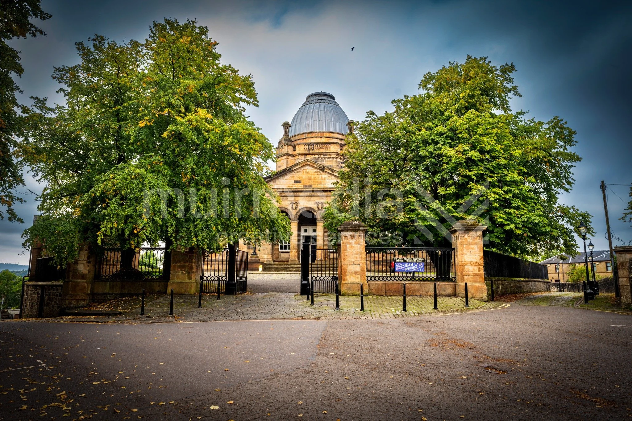 Historic building with dome, flanked by green trees, behind a black wrought iron fence, on a cloudy day.