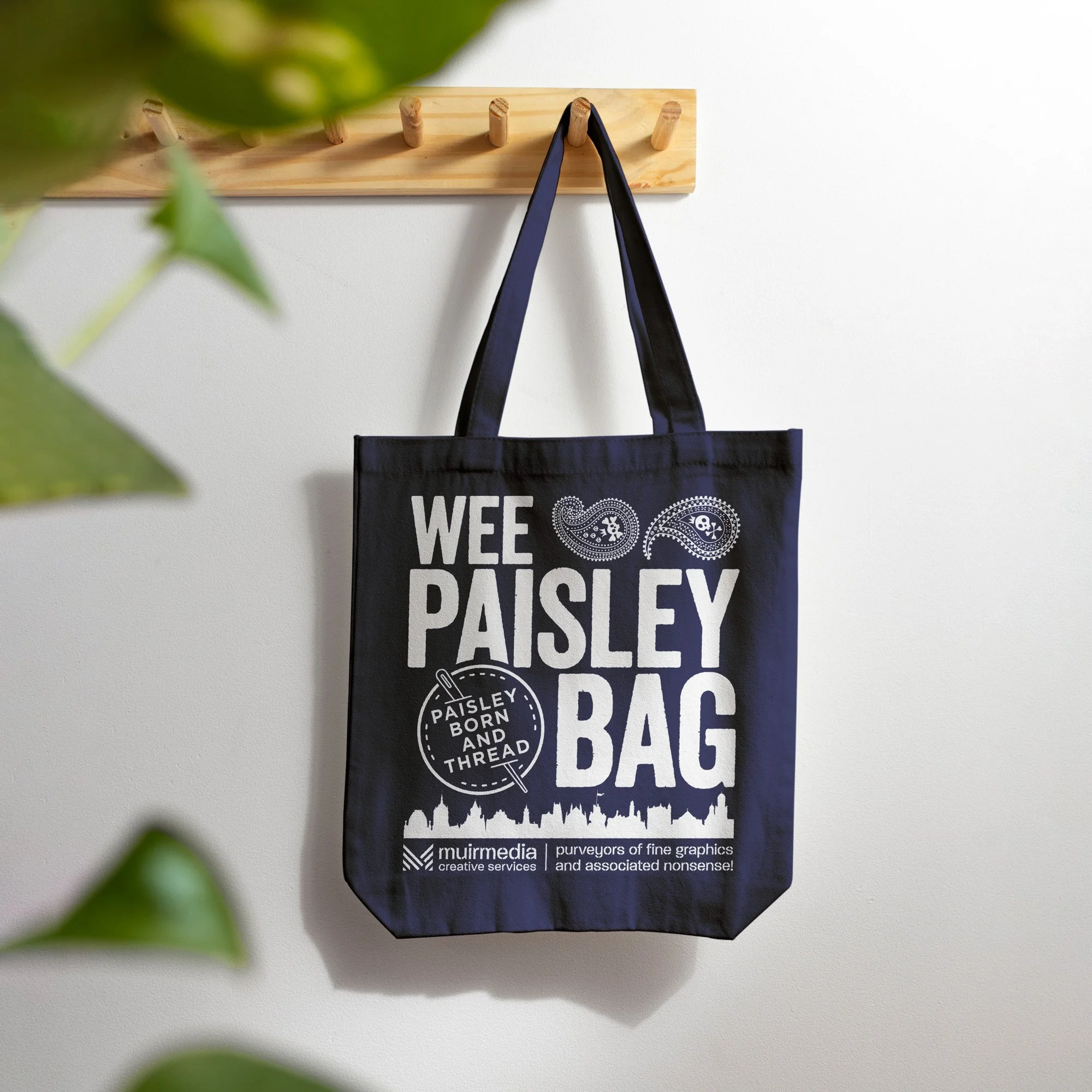 Black tote bag hanging on a wooden wall hook with white text and graphics, reading 'Wee Paisley Bag' and 'Paisley Born and Thread', with logo and cityscape silhouette.