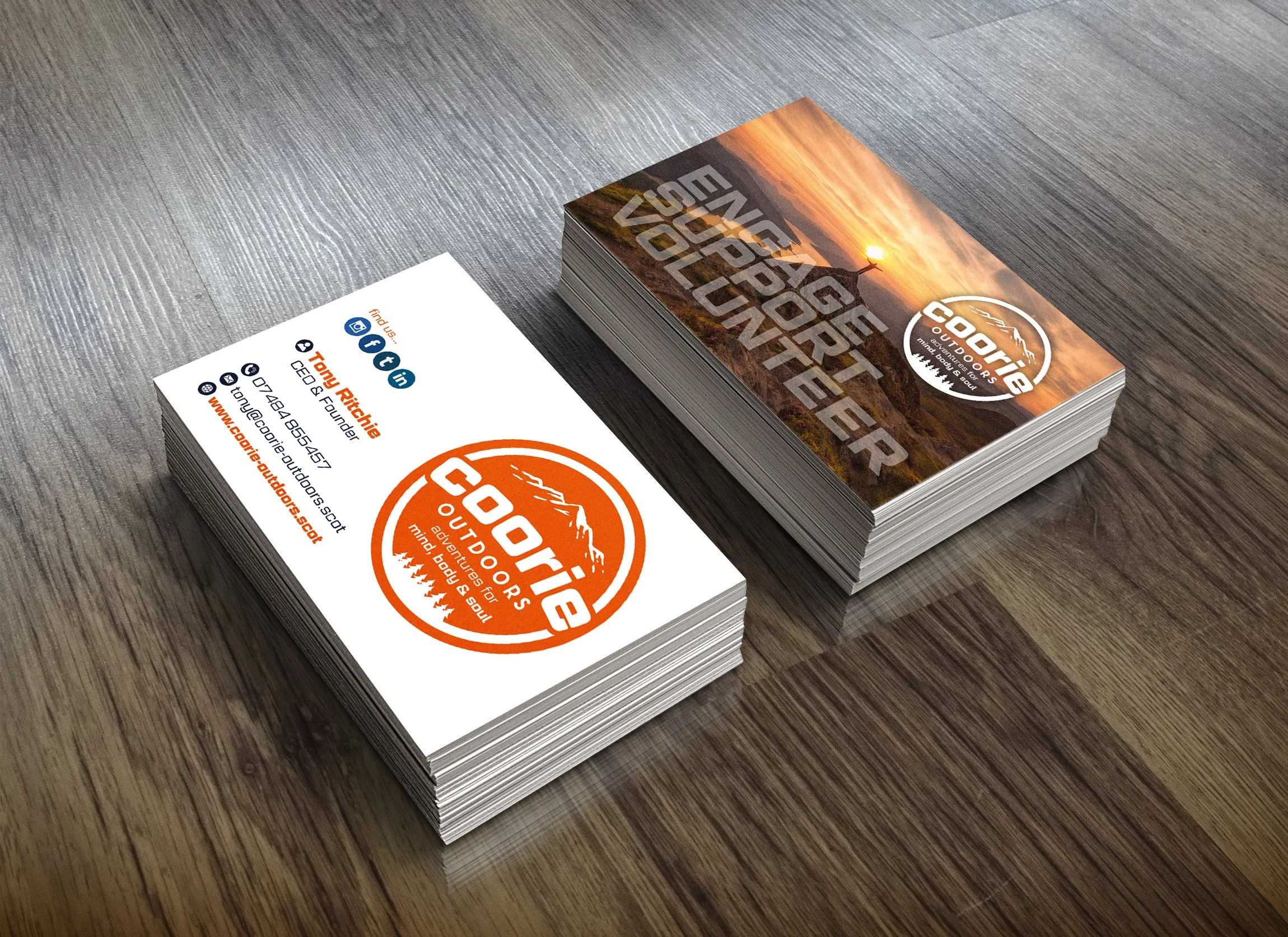 Stack of business cards for CooRe Outdoors featuring a scenic mountain sunset on the front and contact information on the back, placed on a wooden surface.