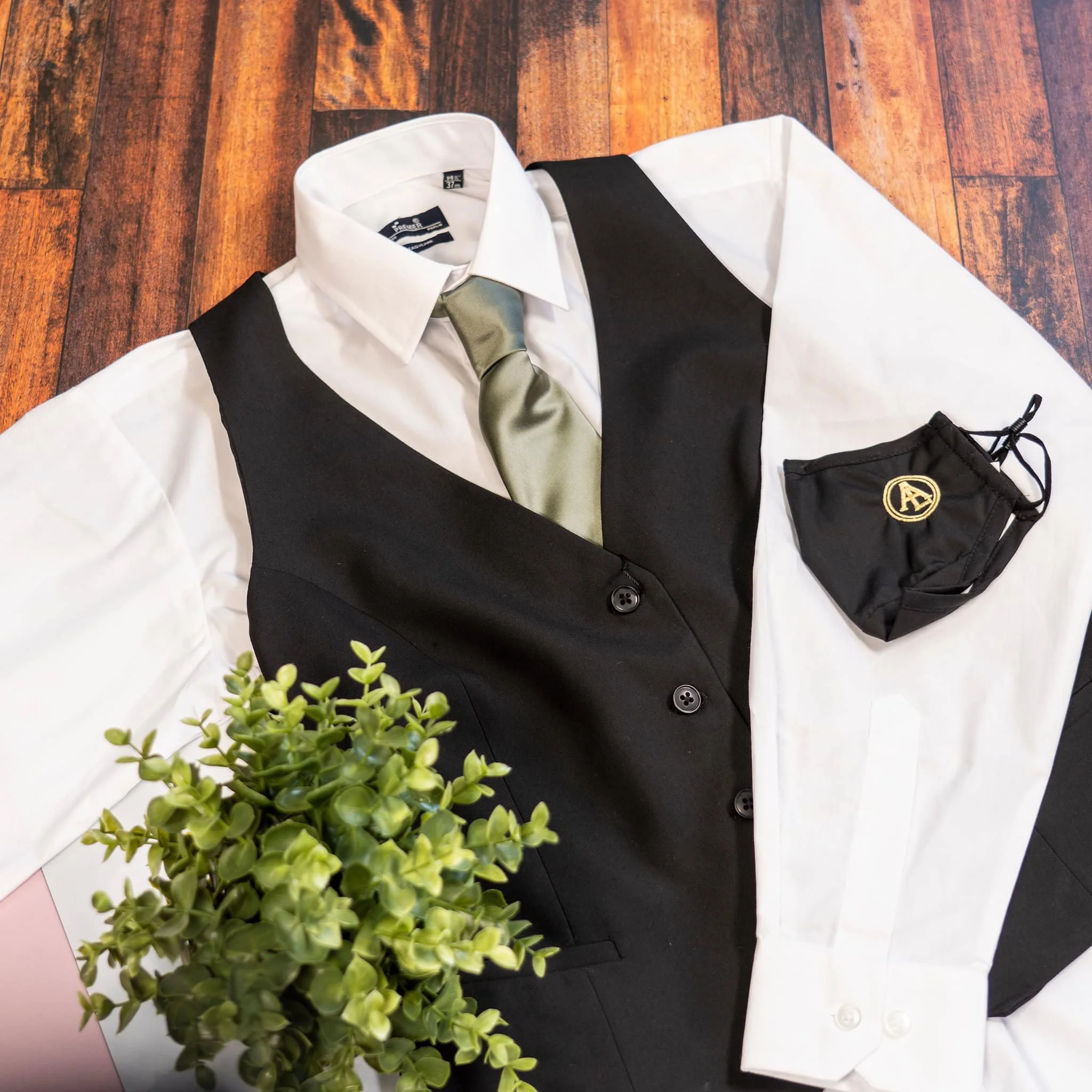 A white dress shirt, a black vest, a light-colored necktie, a black face mask with a gold logo, and a green plant on a wooden surface.