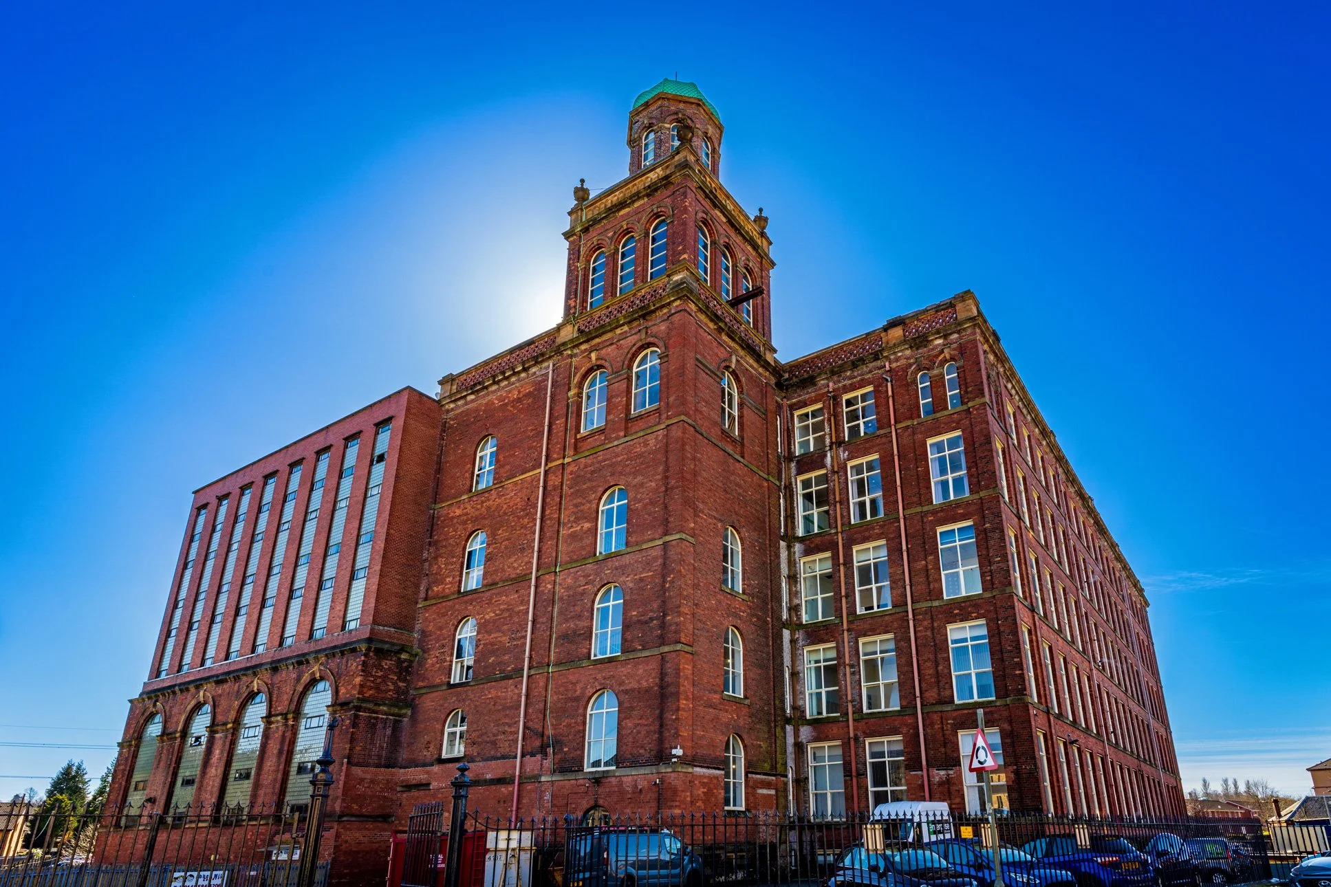 A large red brick building with multiple tall arched windows, a tower with a green dome, and a fence surrounding the property. The sky is clear and blue with the sun shining from behind the tower.