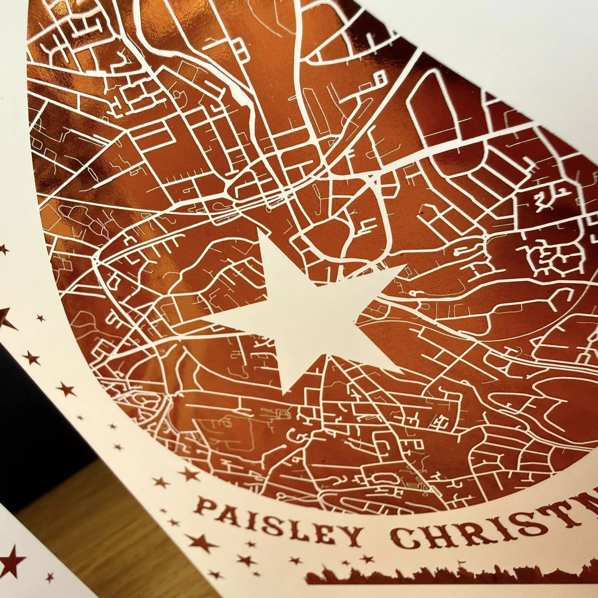 A paper craft of a map of Paisley, featuring streets and roads in white cutout on a brown background, with the name Paisley Christm...