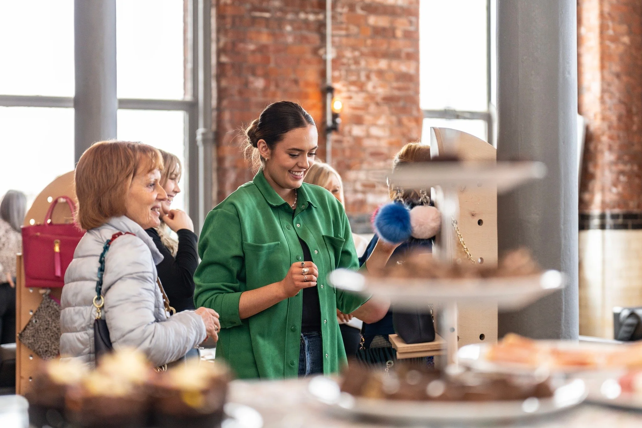 A young woman in a green shirt shopping at a store, smiling as she looks at items on shelves. Two older women and another person are nearby, browsing and chatting in a retail setting with large windows and exposed brick walls.