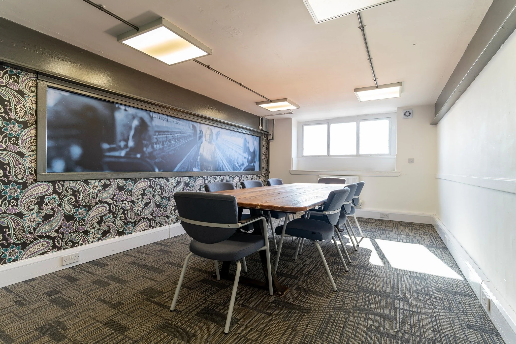 Empty conference room with a wooden table, six chairs, a patterned wallpaper on one wall, a large digital screen, and a window letting in natural light.