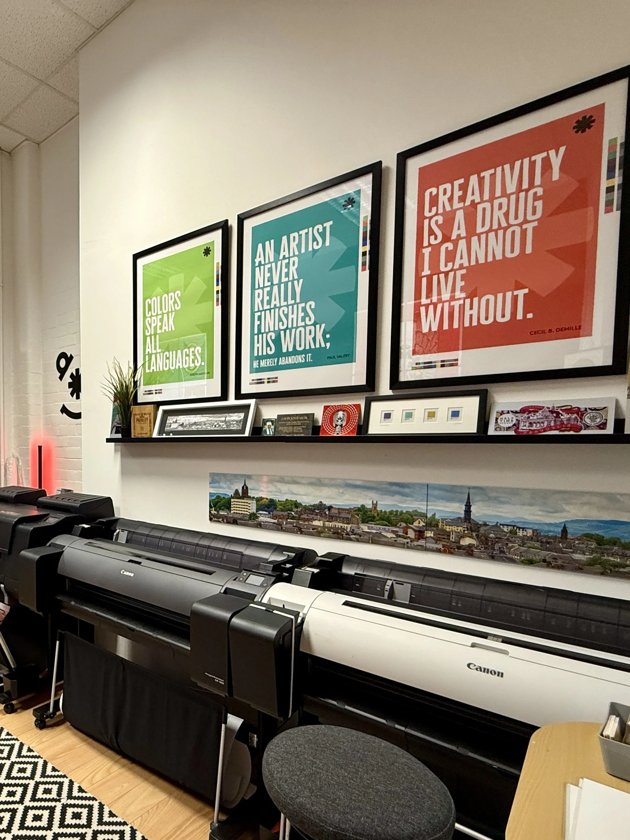 A display of three colorful framed posters on a white wall, with a black shelf holding smaller framed pictures and decorative items below. Large format printers sit on the floor below the shelf, with a patterned rug visible in the foreground.