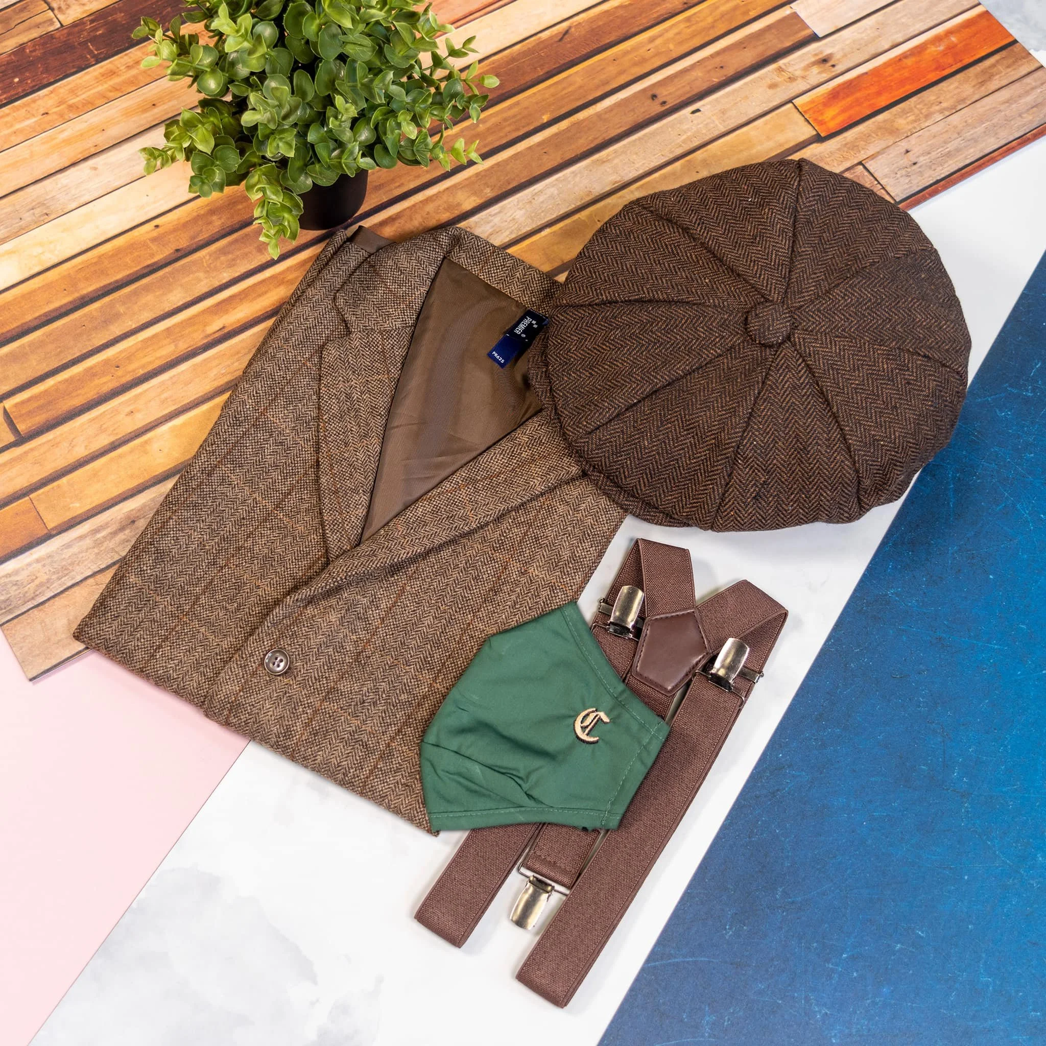 A brown tweed blazer, a brown herringbone flat cap, a green face mask, and brown suspenders on a multicolored surface with wooden plank and marble textures, and a potted plant nearby.