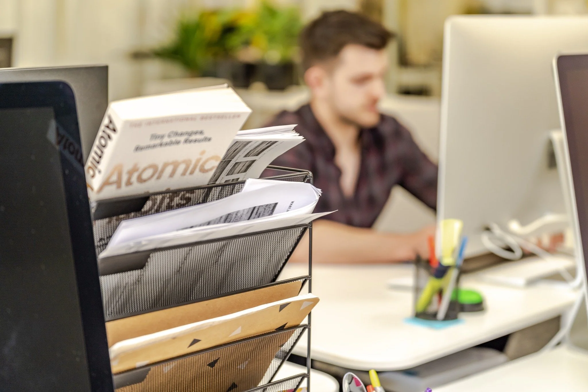 A cluttered office desk with papers, folders, and a book titled 'Atomic' in the foreground. In the background, a man is working at a computer, blurred out.