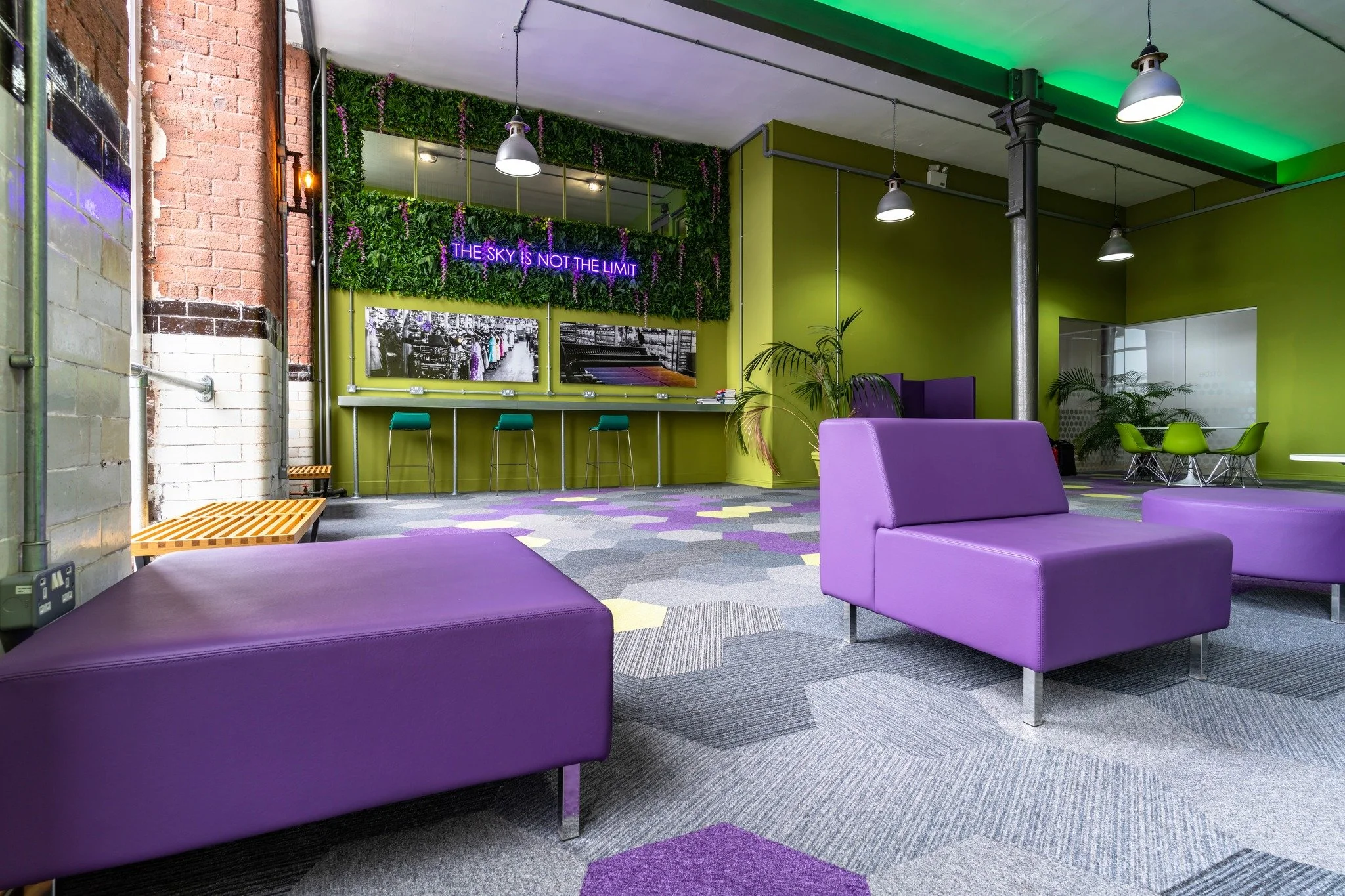 Modern lounge area with purple and green seating, green walls, plants, and wall art, illuminated by ceiling lights.