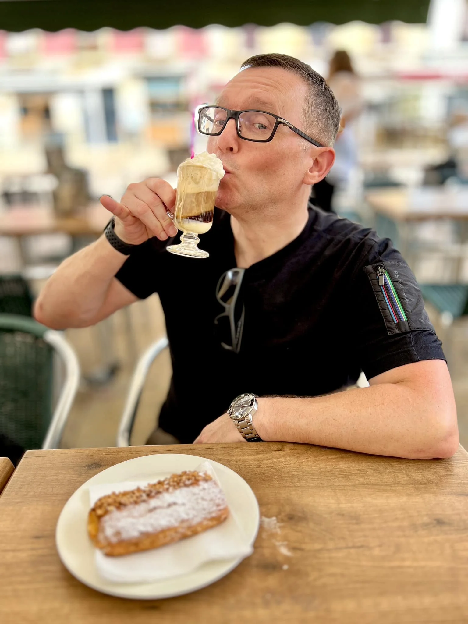 A man in glasses and a black shirt enjoying a layered dessert with whipped cream and chocolate syrup in a glass while seated at a wooden table in a busy cafe or restaurant.