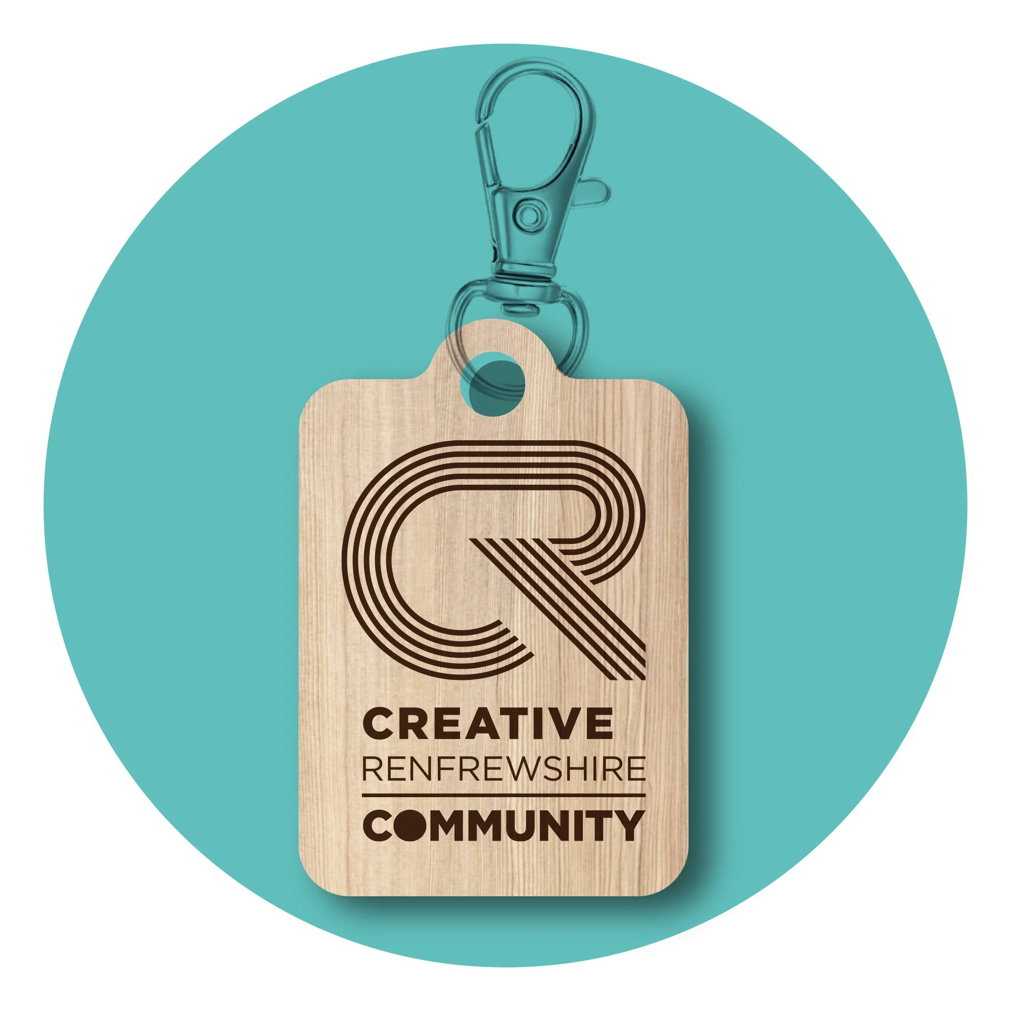 A wooden badge with a black logo and text, attached to a clear plastic clip and keychain against a teal circular background. The badge displays the words "Creative Renfrewshire Community".