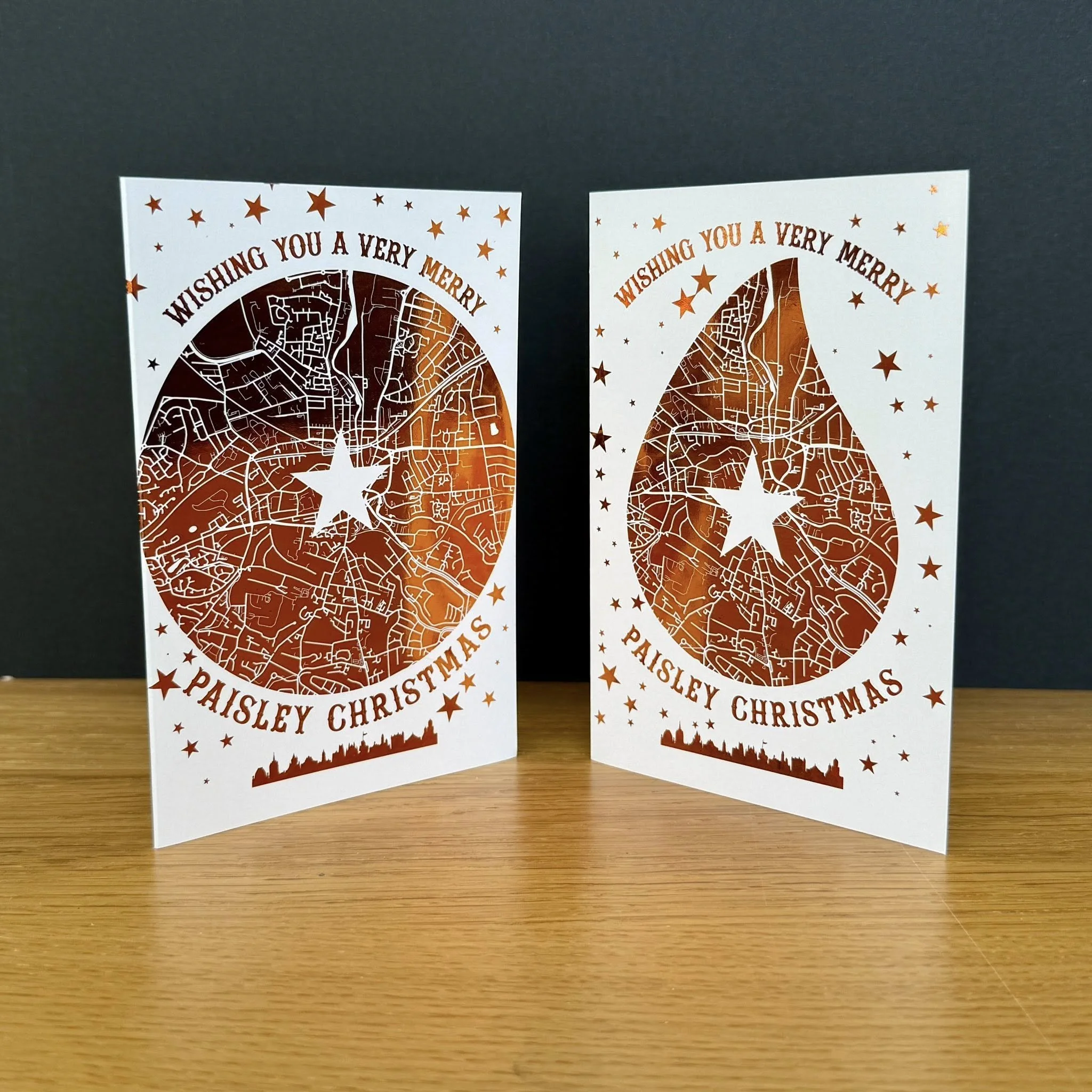 Two Christmas cards with cut-out designs of Paisley featuring a map pattern and a star in the center. The cards are decorated with stars and the words 'Wishing You a Very Merry Paisley Christmas' in brown lettering.