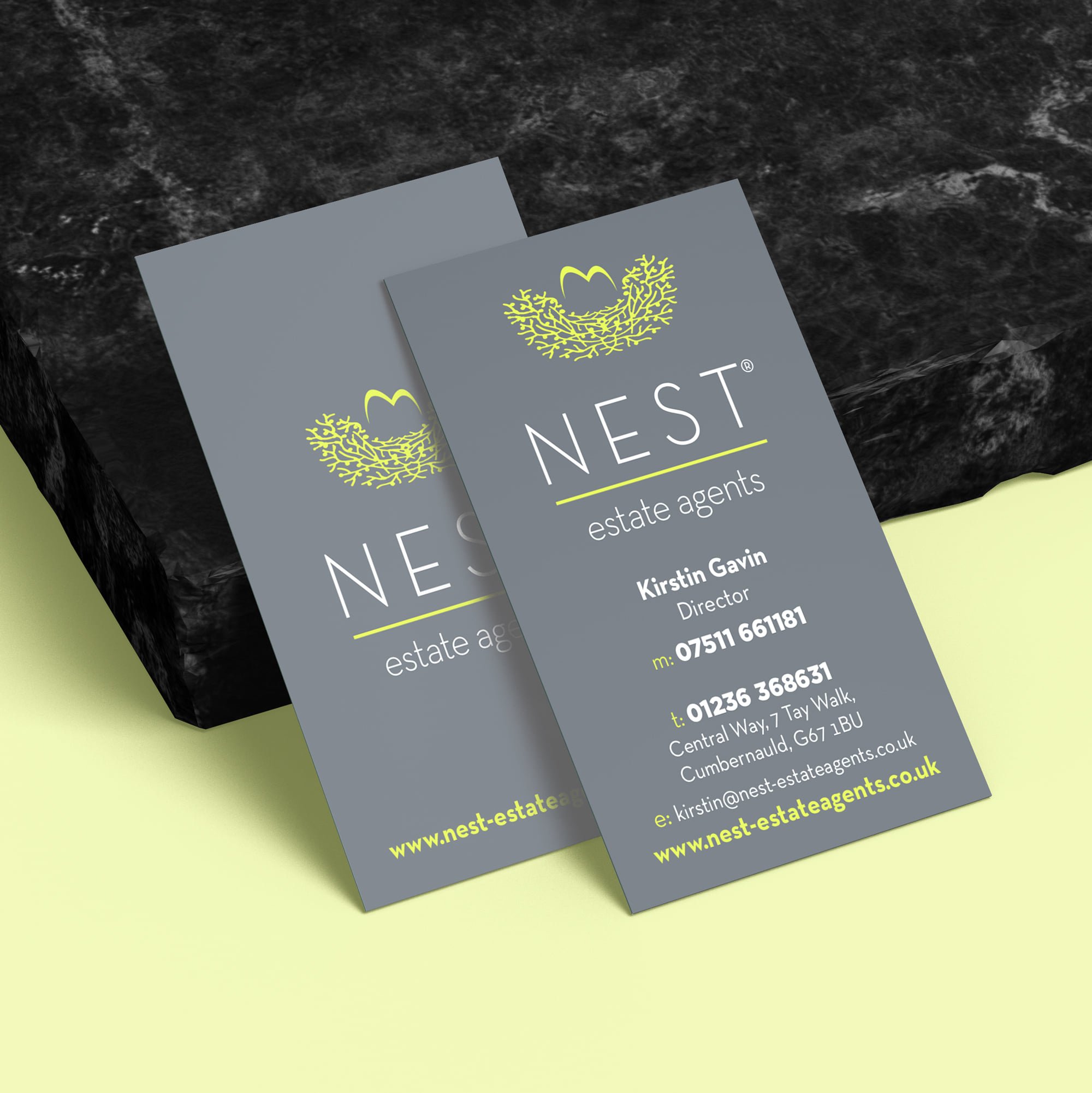 Close-up of a gray business card for NEST estate agents with yellow and white text, beside a black textured surface on a light background.