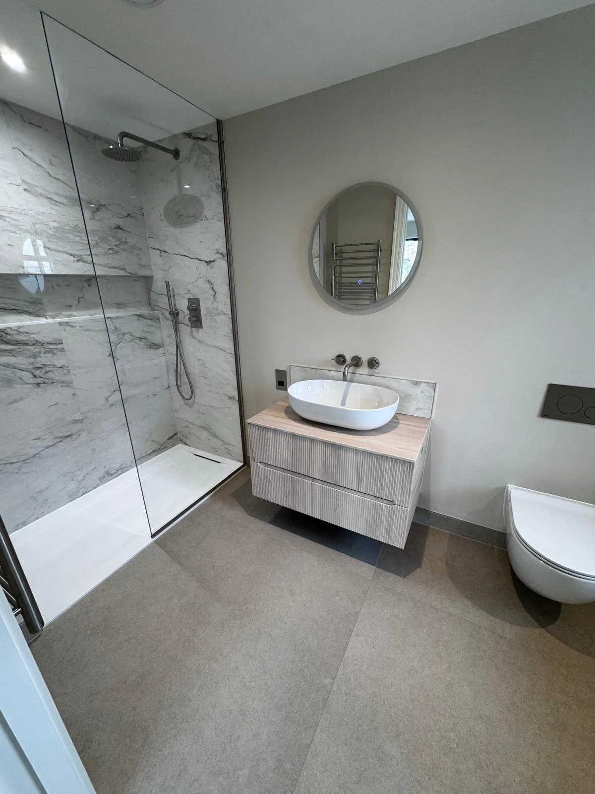 Modern bathroom with a marble-tiled shower enclosure, a round mirror above a wooden vanity with a bowl sink. Neutral tones create a clean, minimalist look.