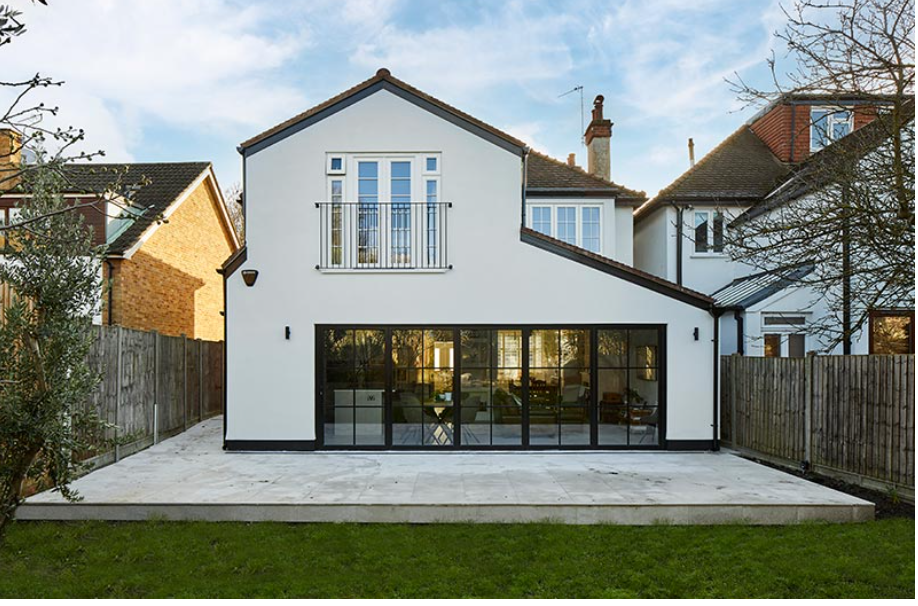 Two-story white house with a gable roof, featuring a small balcony and large glass sliding doors on the lower level. The sky is clear and blue.