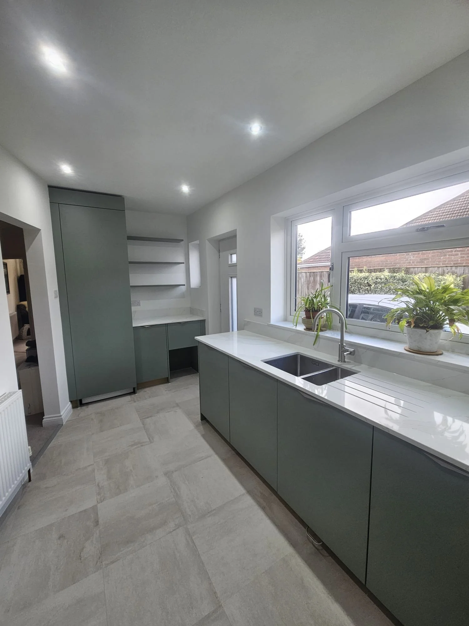 Modern kitchen with sleek gray cabinets and white countertops. Large window with potted plants provides natural light. Minimalist design, calm ambiance.