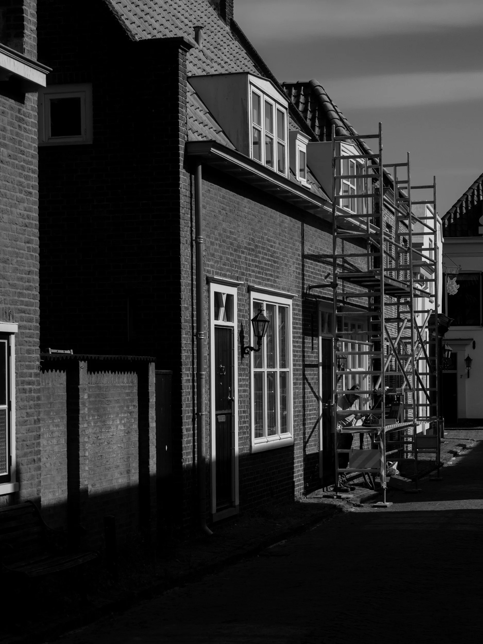 The image is a black and white photo of a brick building with a row of windows and a lamp. Scaffolding is set up on the side, casting shadows.