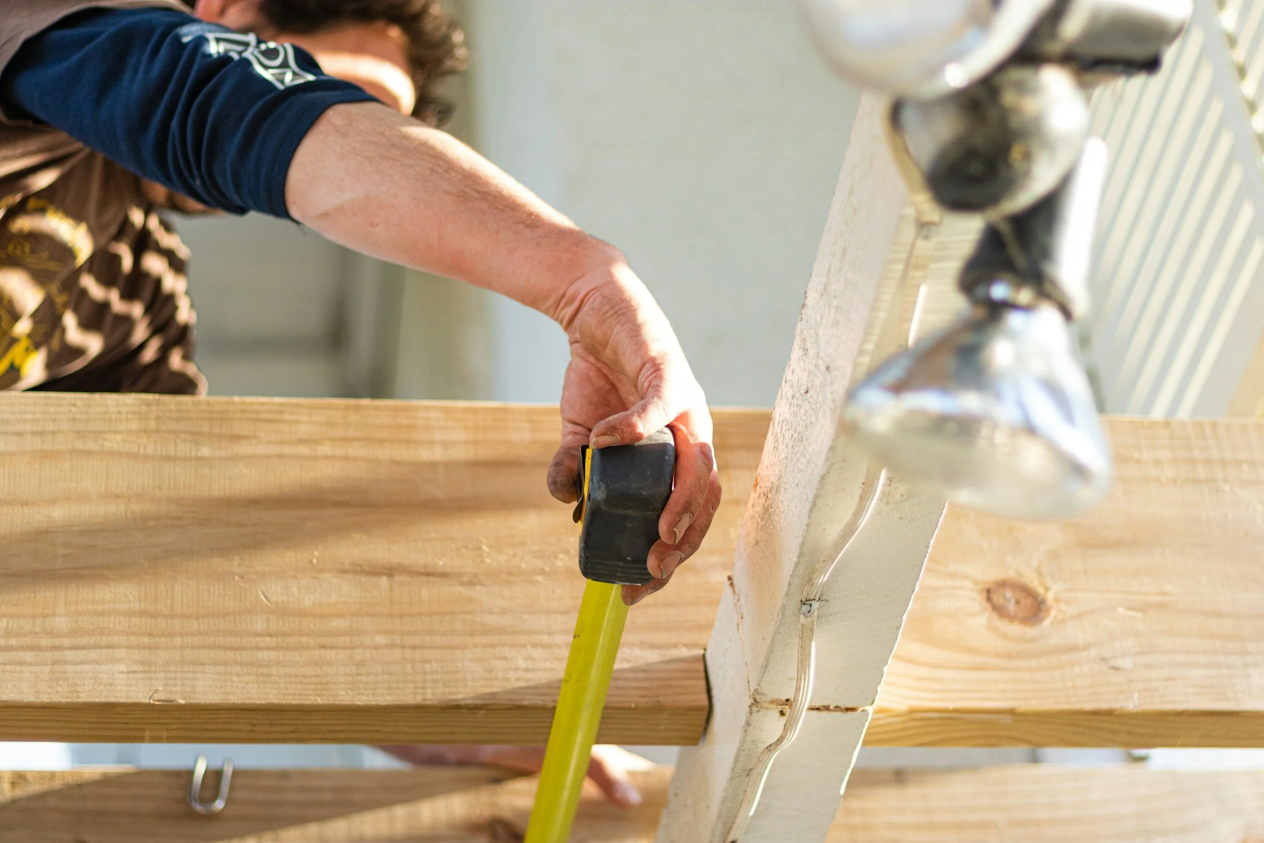 A person measures a wooden plank with a yellow tape measure. The scene conveys a focus on DIY woodworking, with light streaming in, creating a warm tone.