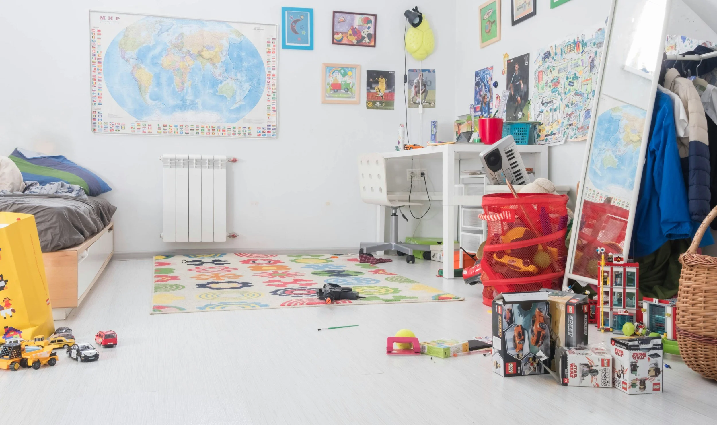 Child's bedroom with bed, toy cars, colorful rug, wall map, and sports posters, cluttered with toys, boxes, and clothing.
