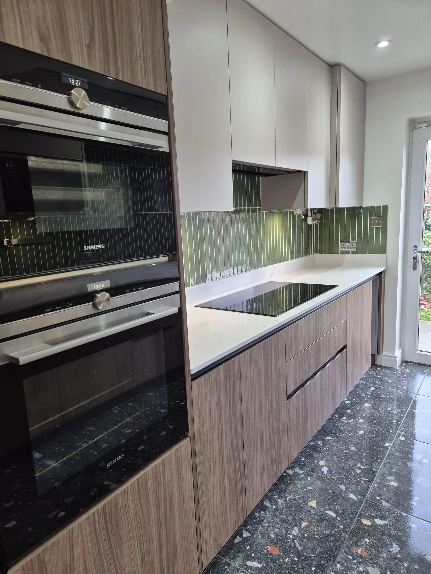 Modern kitchen with sleek wood cabinetry, white countertops, and green tile backsplash. Features built-in ovens, electric stove, and terrazzo flooring.