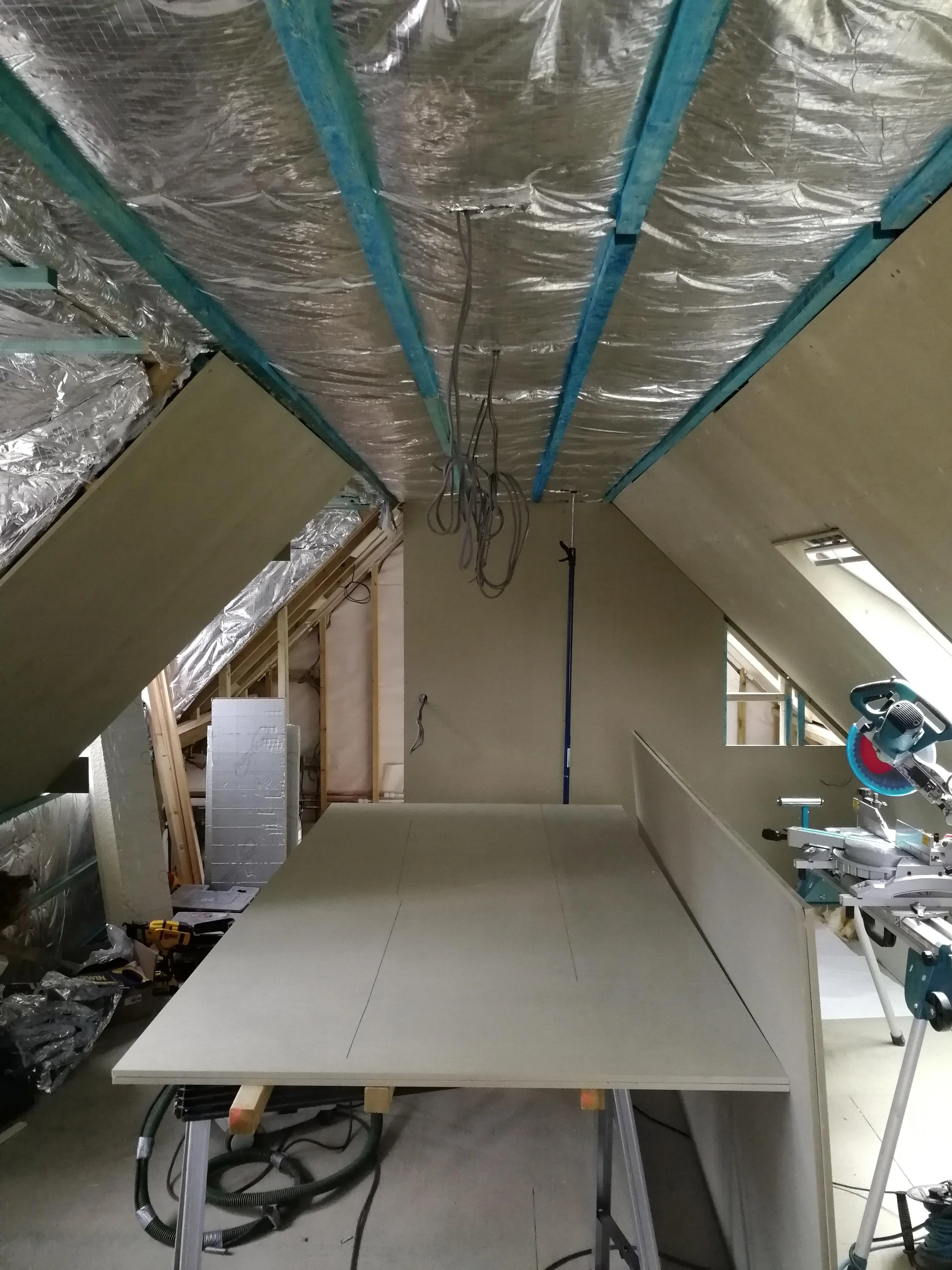 Attic under renovation with exposed insulation and wiring on the ceiling. Drywall sheets are laid out on sawhorses, alongside construction tools.
