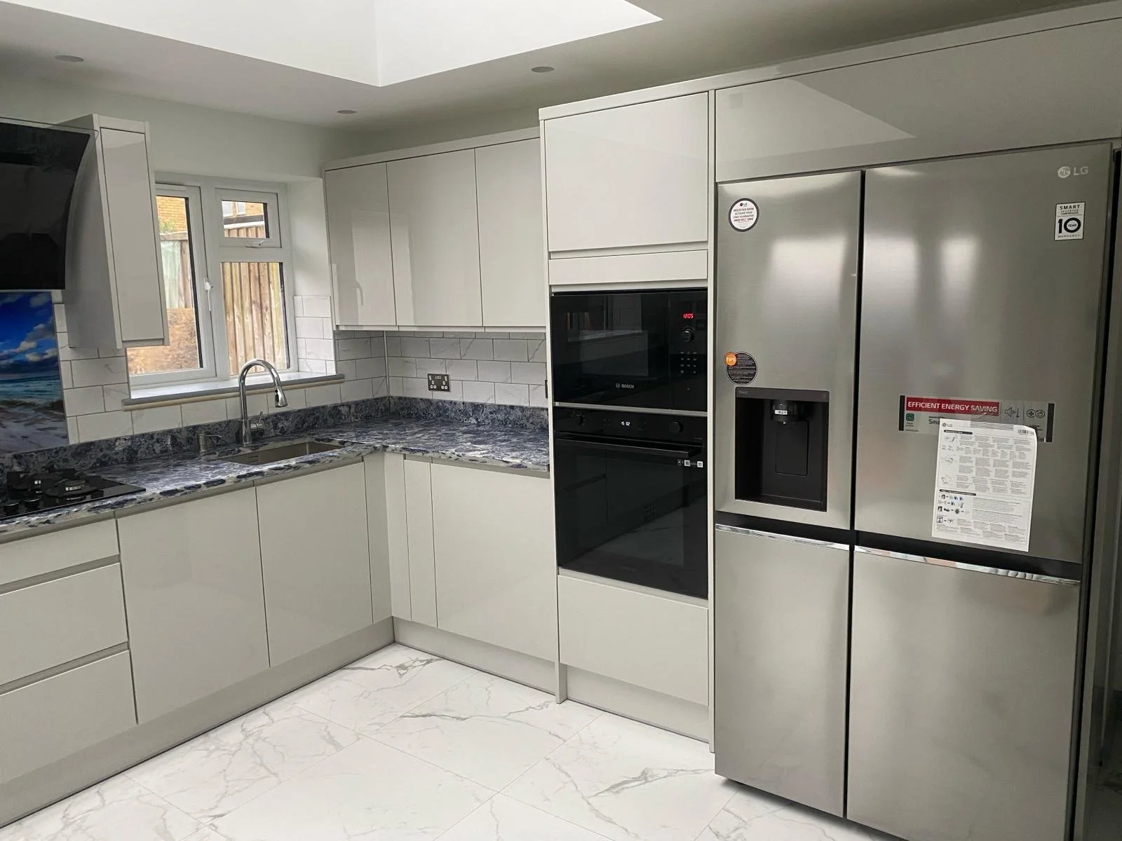 Modern kitchen with gray cabinets, marble countertops, and stainless steel appliances, including a double-door fridge. Bright lighting creates a sleek, clean look