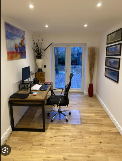 Cozy home office with a wooden desk, computer, and chair. Glass doors overlook a garden, with art and photos on white walls. Calm, organized ambiance.