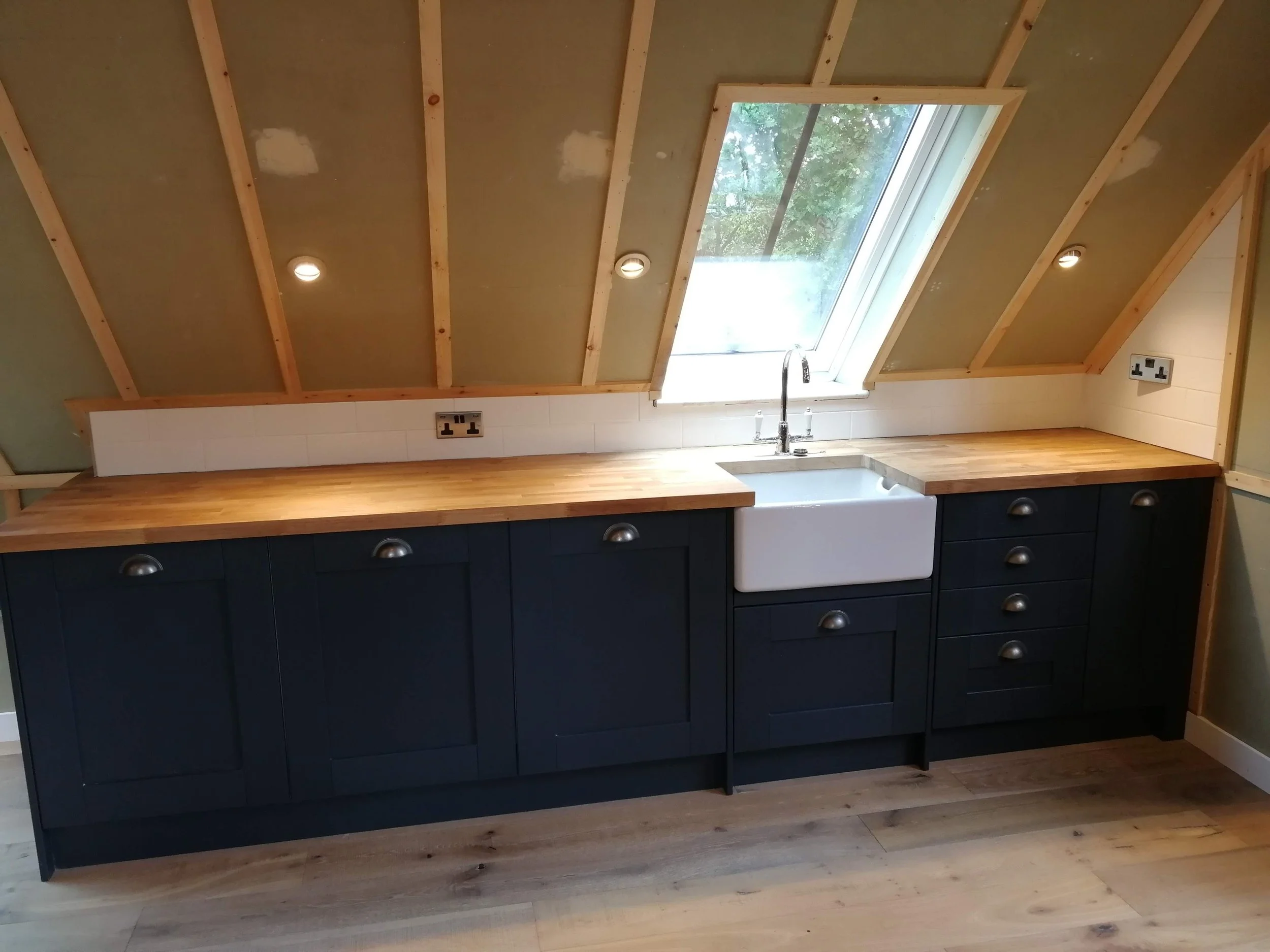 Attic kitchen with sloped wooden beams, pendant lights, black cabinets, wood countertops, and a farmhouse sink. Bright and minimalist design.