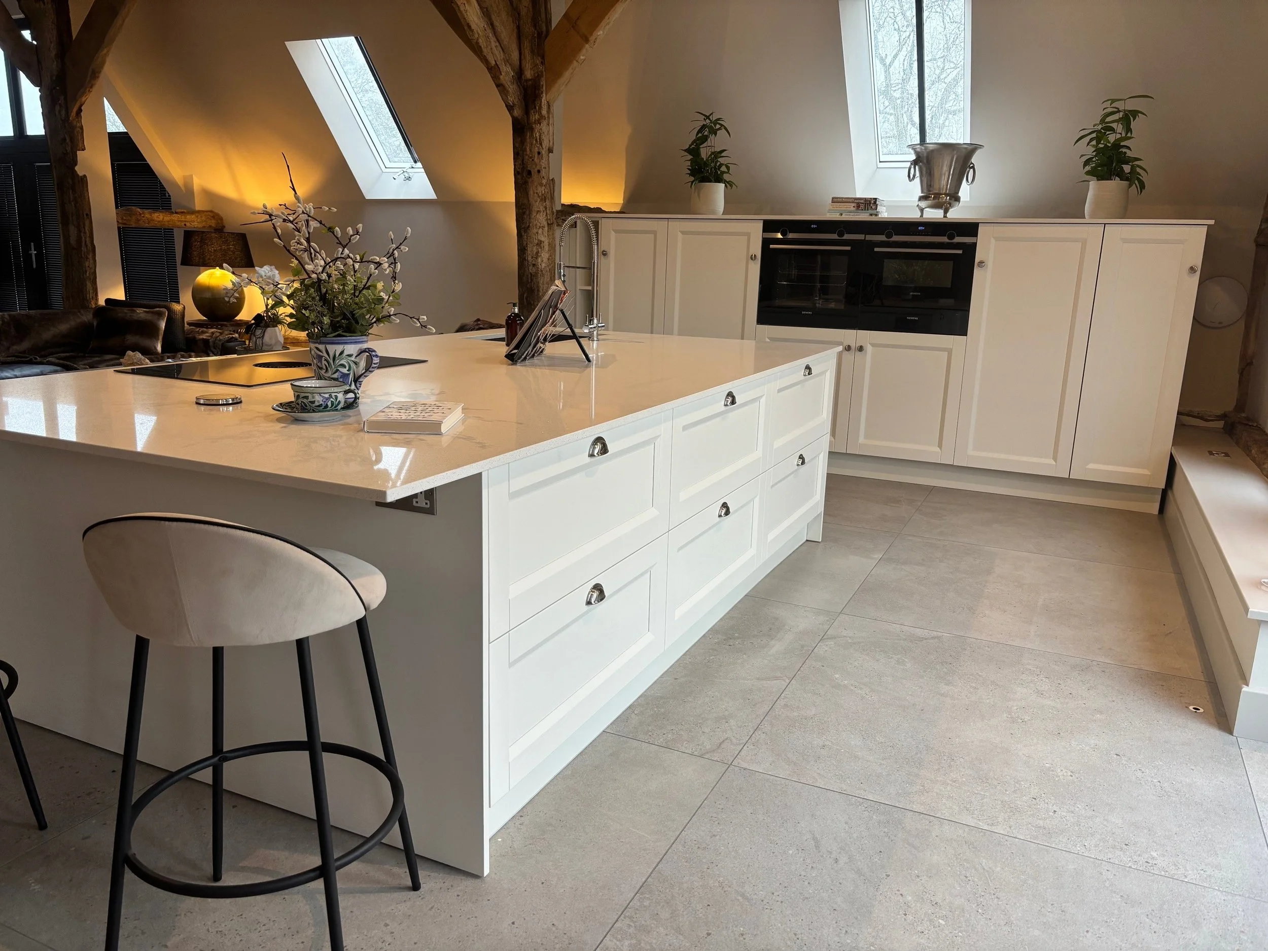 Modern rustic kitchen with a white island, integrated oven, and wooden beams. Soft lighting creates a warm, inviting atmosphere. Skylight and potted plants add freshness.