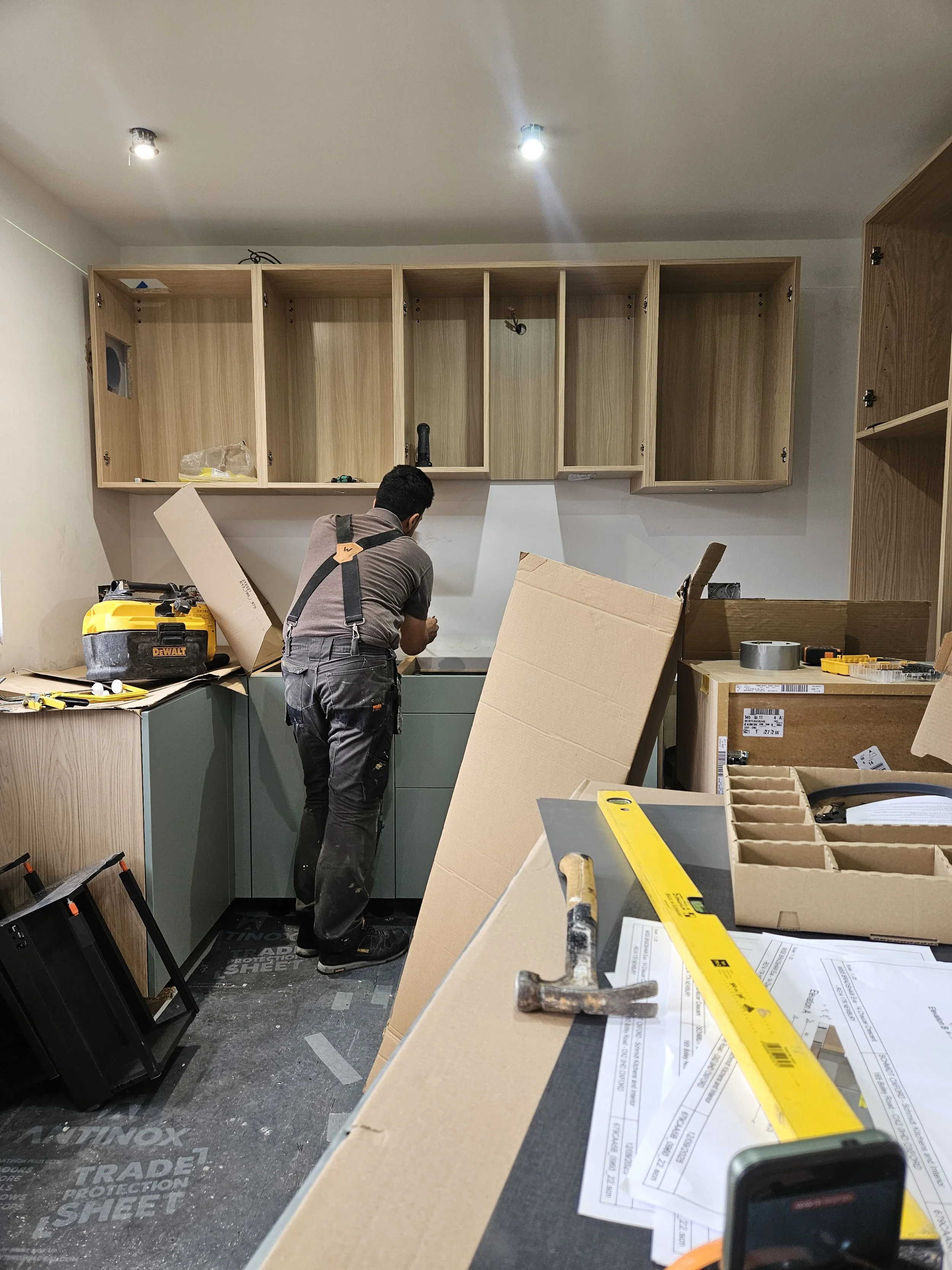 A man in overalls works on a kitchen installation. The room is cluttered with tools, cardboard boxes, and wooden cabinets, conveying a work-in-progress feel.