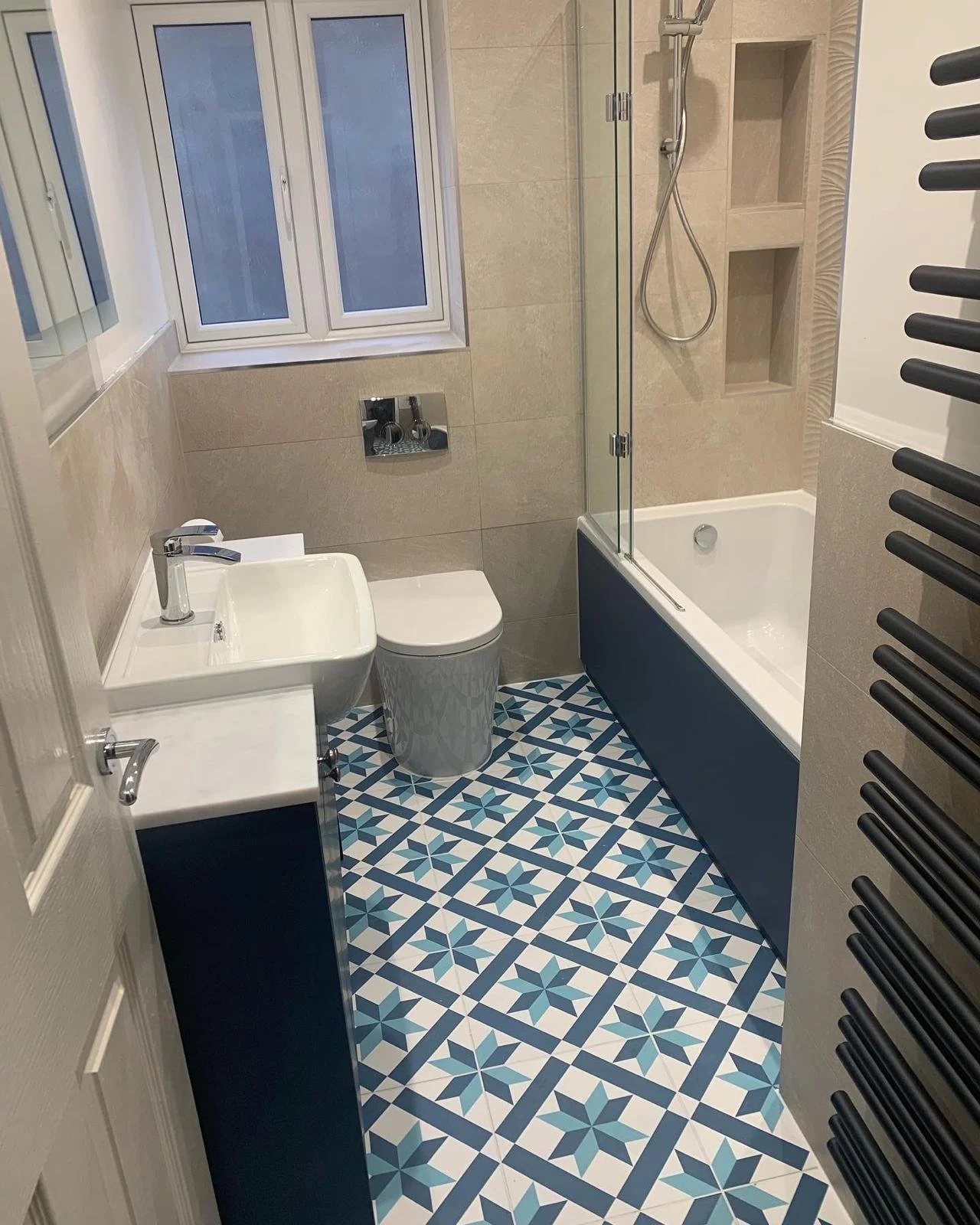 A small, modern bathroom features geometric blue and white floor tiles, a white sink and toilet, a bathtub with a glass shower screen, and beige walls.
