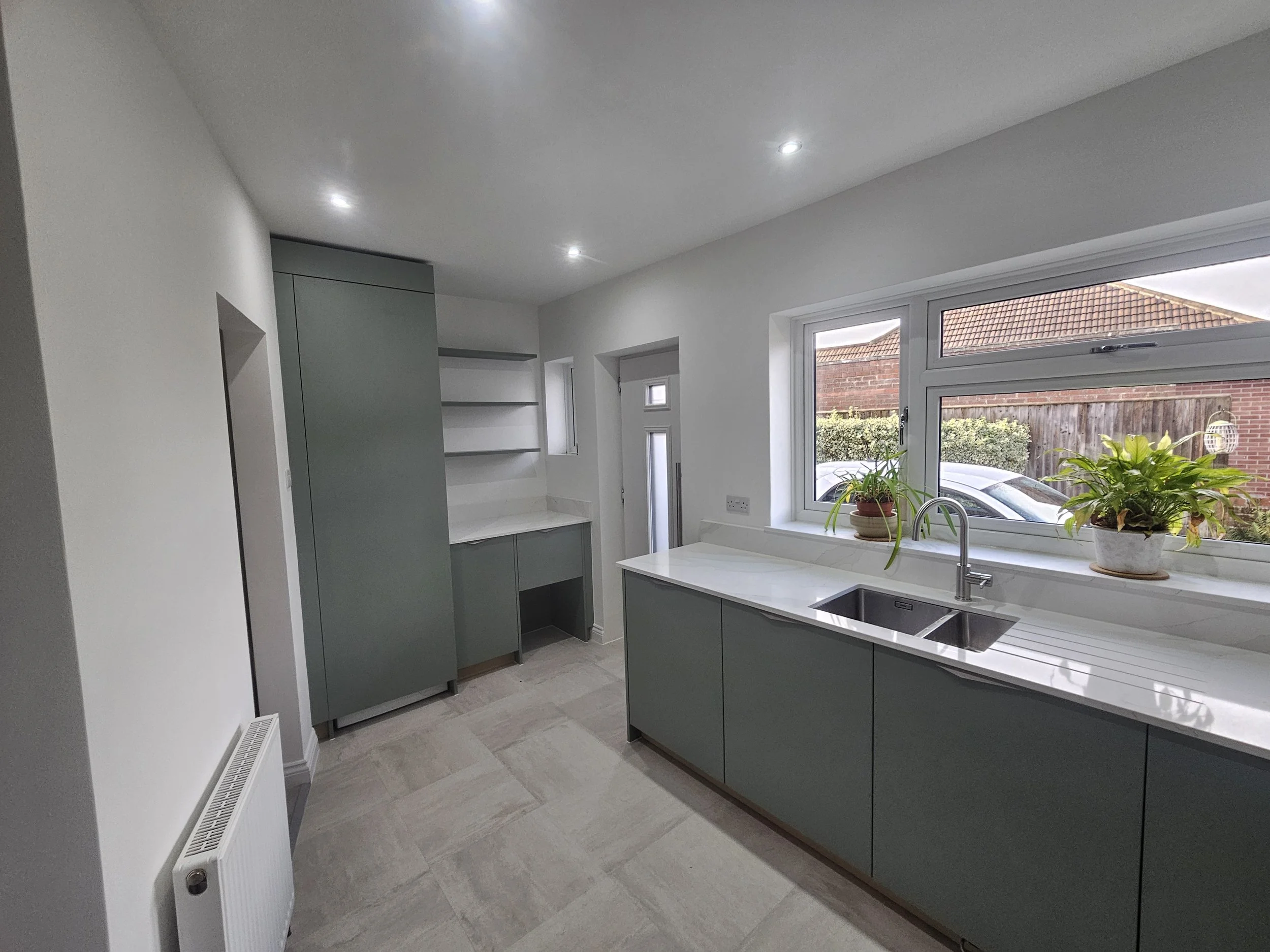 Modern kitchen with green cabinets, marble countertops, and stainless steel appliances. Large window with potted plants adds natural light and warmth.