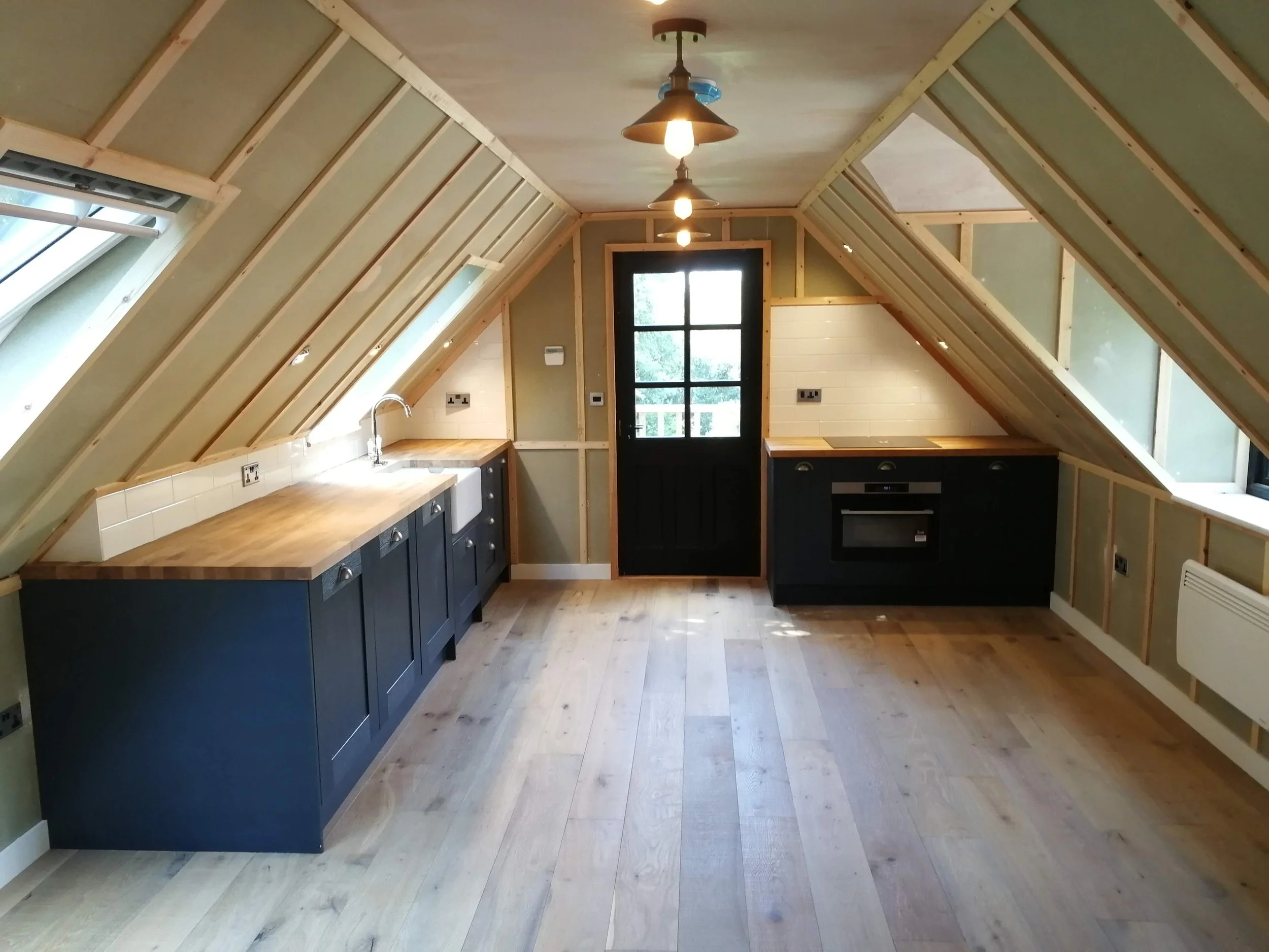 Attic kitchen with sloped wooden beams, pendant lights, black cabinets, wood countertops, and a farmhouse sink. Bright and minimalist design.