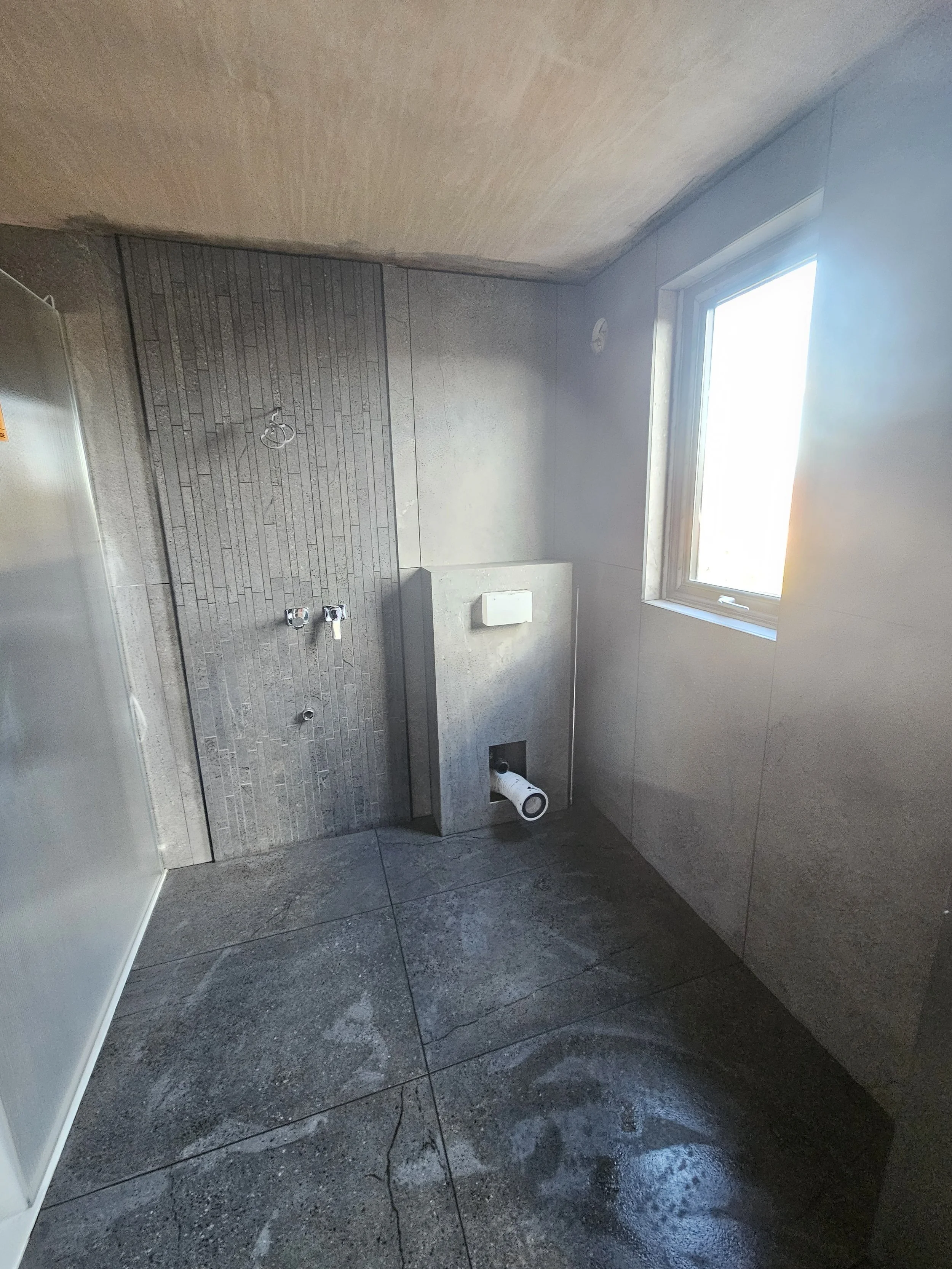 Minimalist bathroom under construction with gray stone tiles, a rough concrete wall, exposed plumbing, and a window letting in soft natural light.