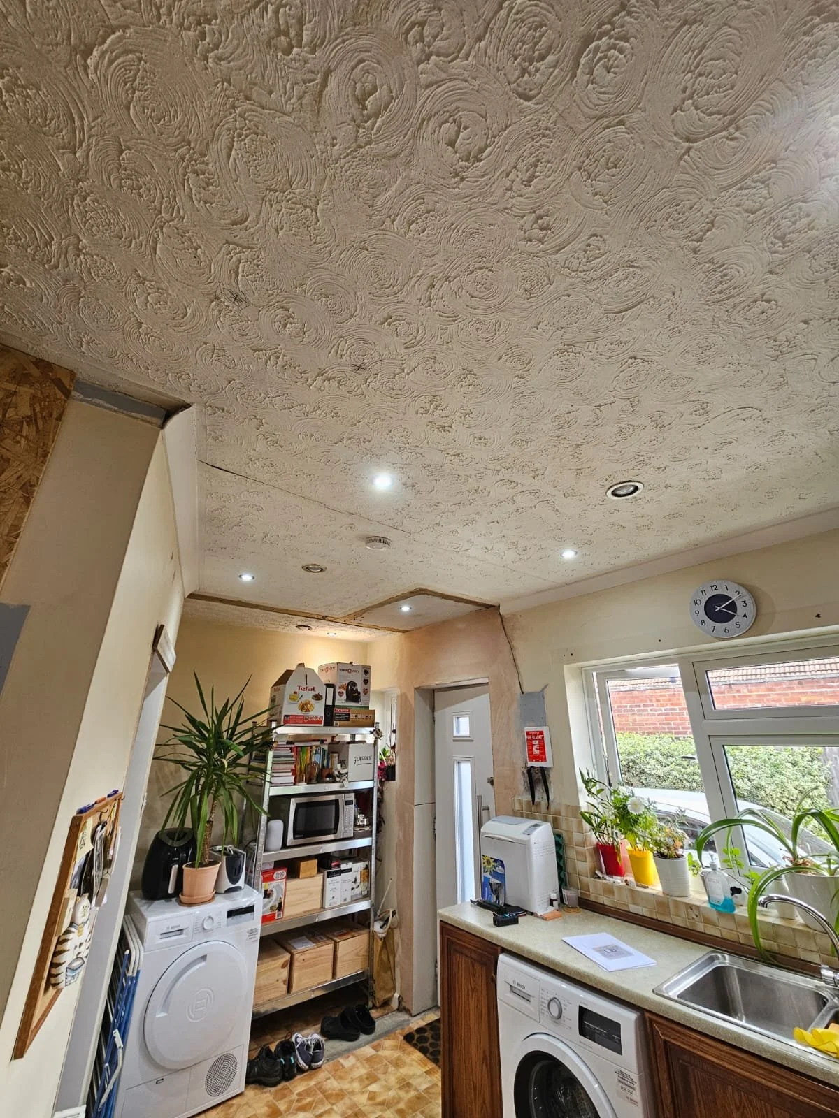 A cozy kitchen with a textured, white ceiling and recessed lighting. A plant, appliances, and colorful potted herbs line countertops near a window.