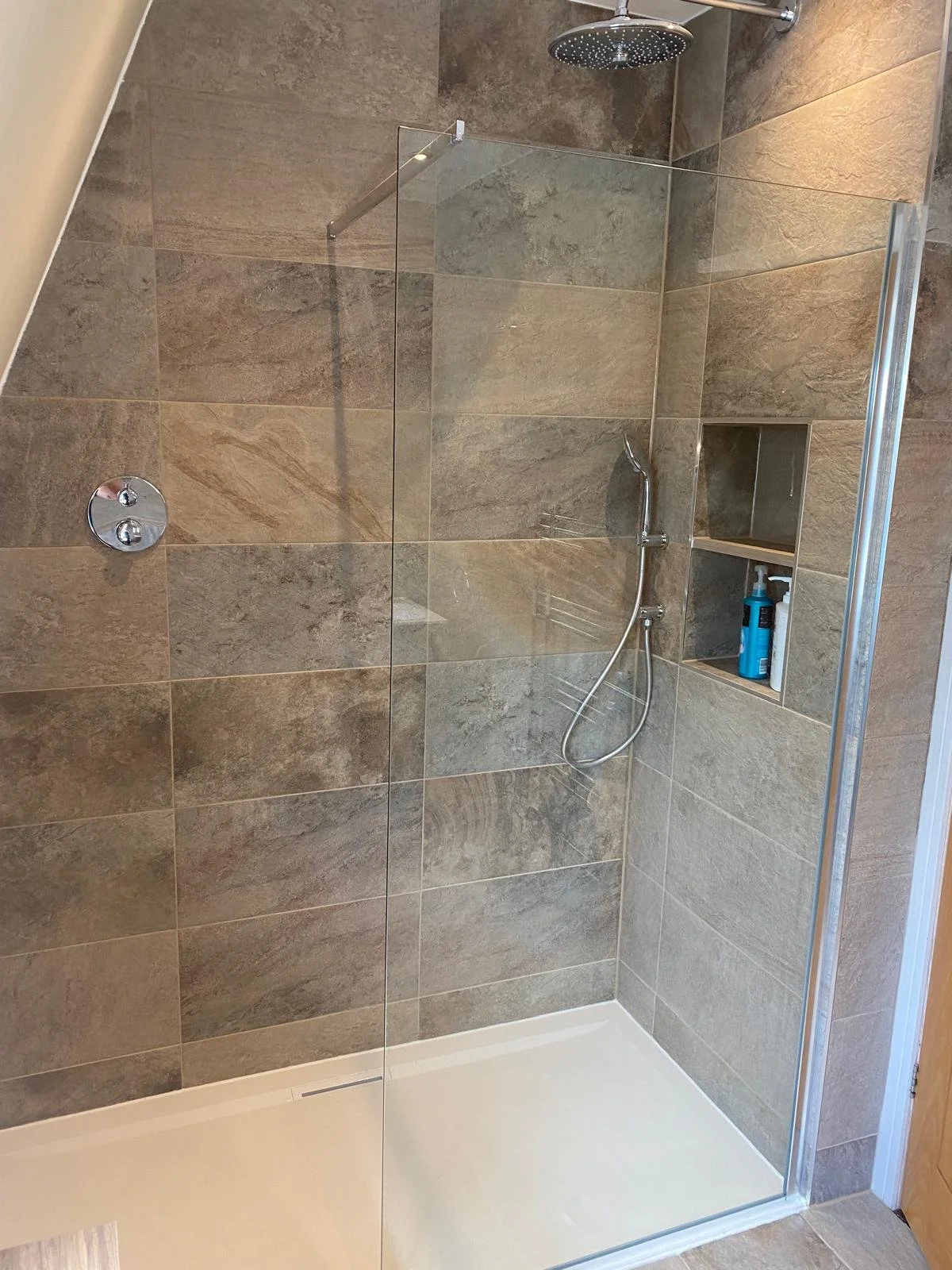 A modern shower with beige stone tiles, a glass panel, and a rainfall showerhead. Built-in shelf holds toiletries, creating a clean and serene atmosphere.