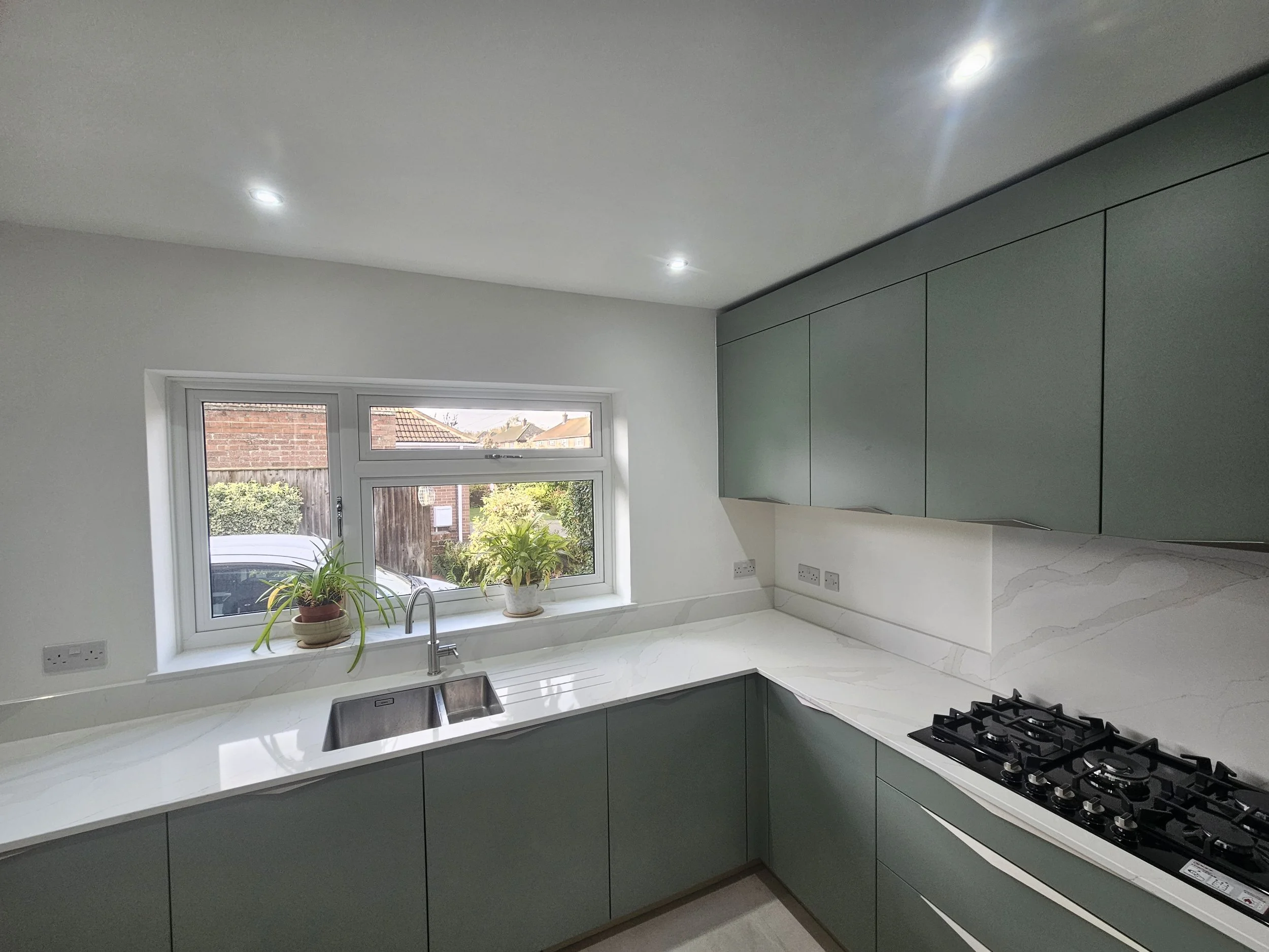 Modern kitchen with sleek green cabinets and a white marble countertop. A stainless steel sink is under a window decorated with potted plants. Cozy and bright.
