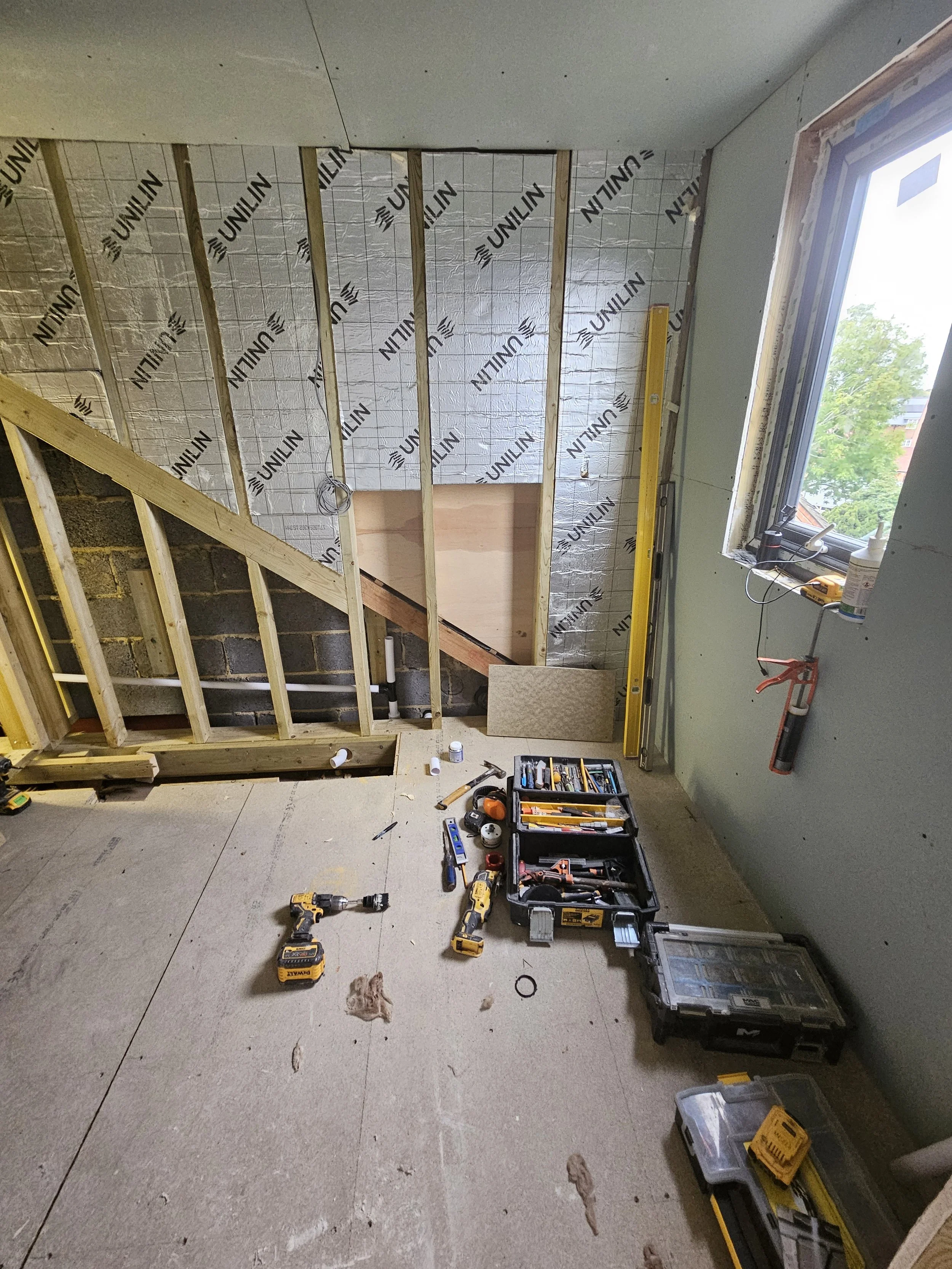 Room under construction with exposed plywood walls and insulation. Tools scattered on the floor include a drill and toolbox. A window offers natural light.