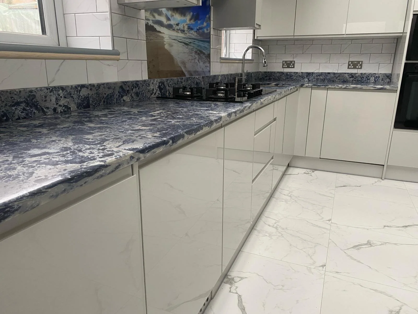 Modern kitchen with sleek white cabinets, blue marbled countertops, and tiled walls. It features a stainless steel sink on a shiny floor. Coastal artwork adds a calm feel.