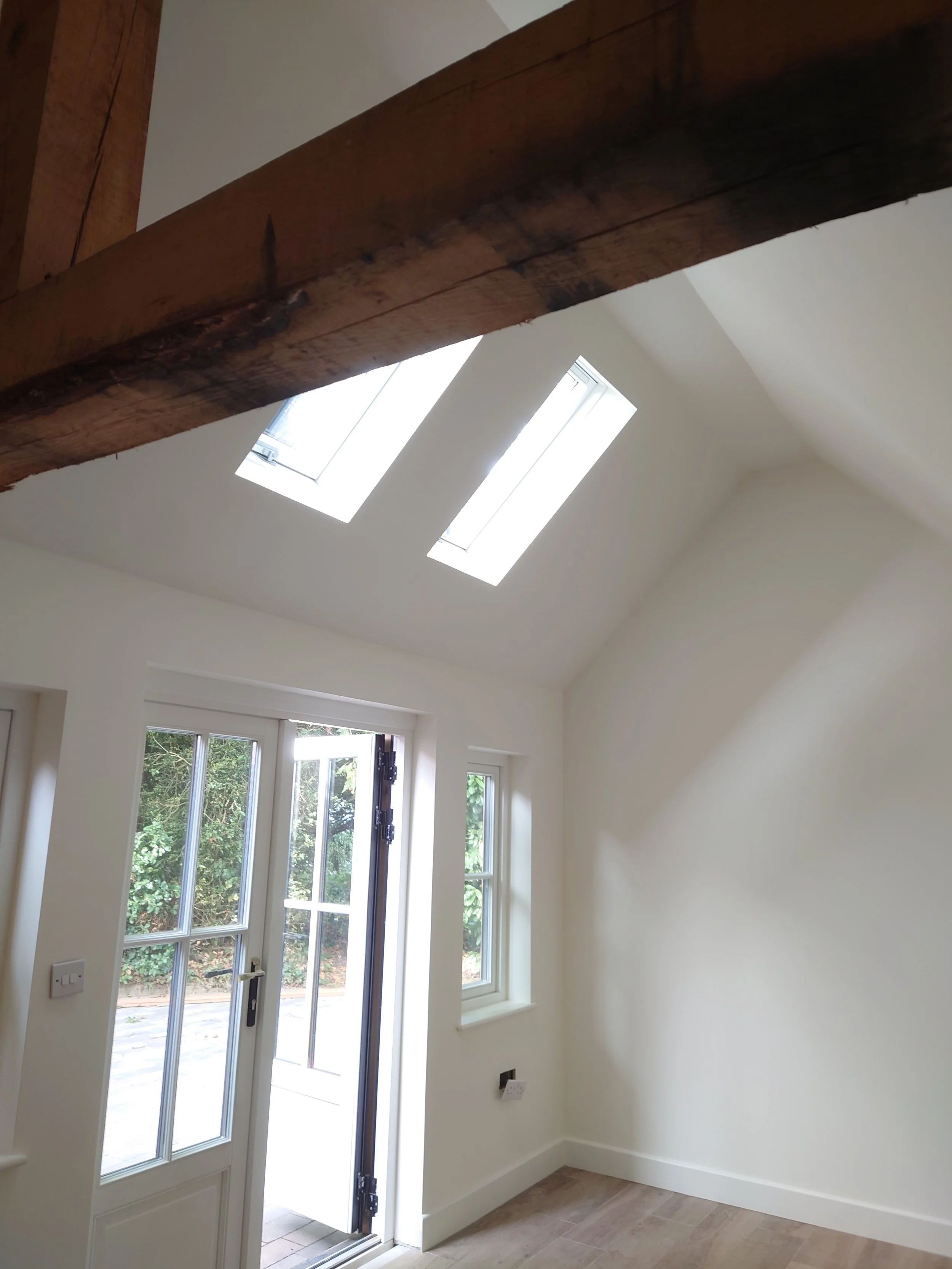 Bright room with white walls, wooden floor, and exposed beam. Sunlight streams through two skylights and large glass doors, creating a serene, airy feel.