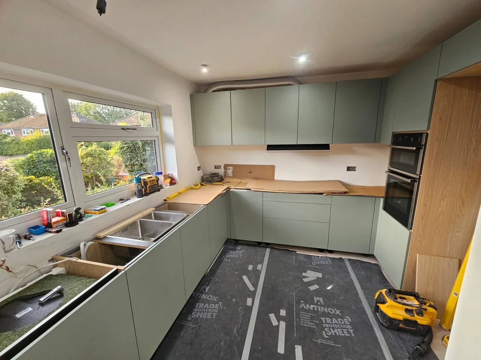 Modern kitchen under renovation with light green cabinets, unfinished countertops, and a double sink. Tools and materials are scattered, conveying progress.