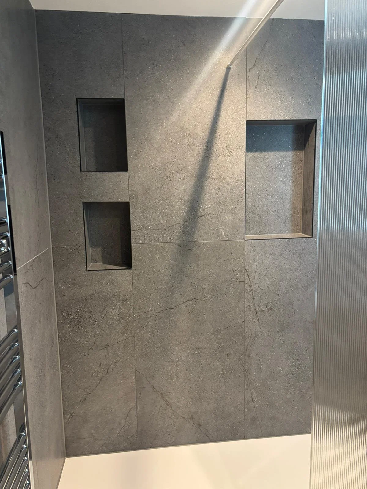 Textured gray tiled wall with built-in niches, casting subtle shadows. A smooth, sleek design featuring a chrome towel radiator on the left.